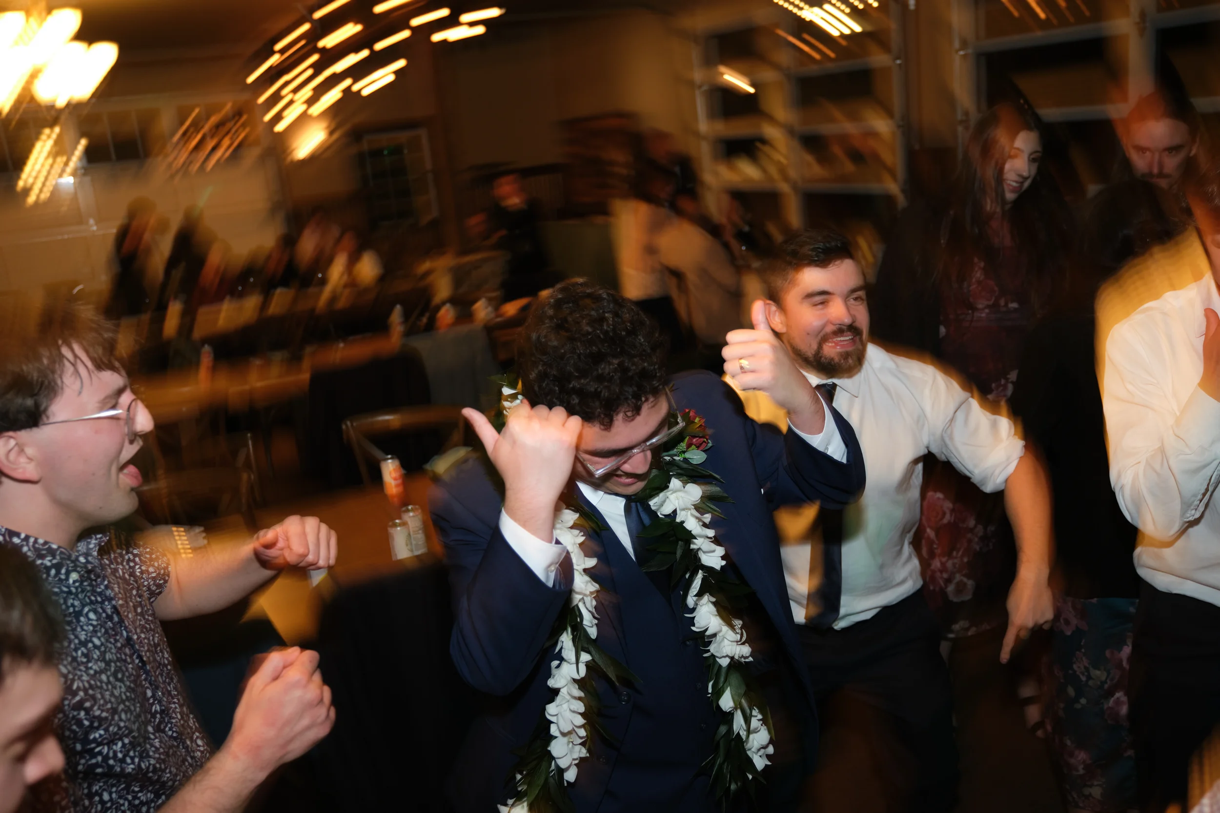 People celebrating at a wedding reception, smiling and dancing, with some wearing floral leis. The background shows a warm, well-lit indoor venue.