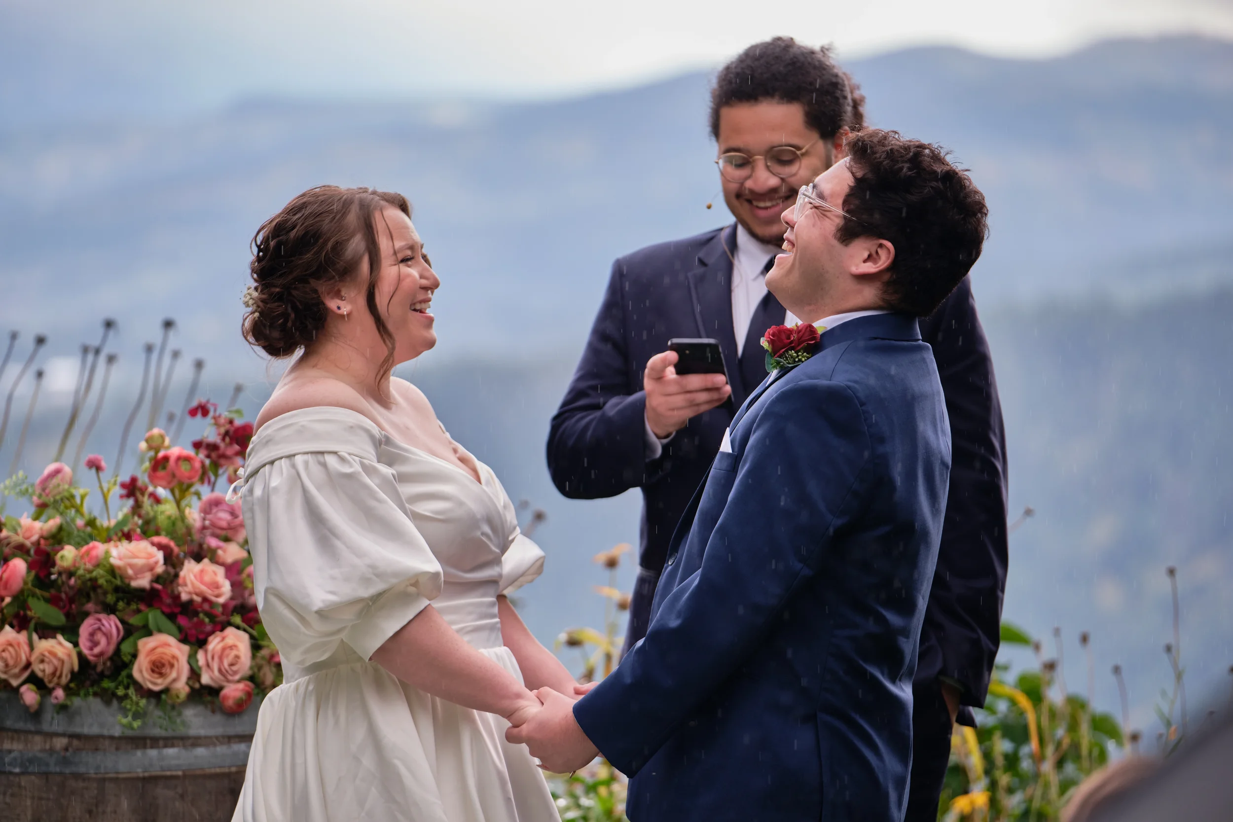A couple getting married outdoors, standing close and holding hands, with a officiant smiling in between, surrounded by flowers and a cloudy sky.