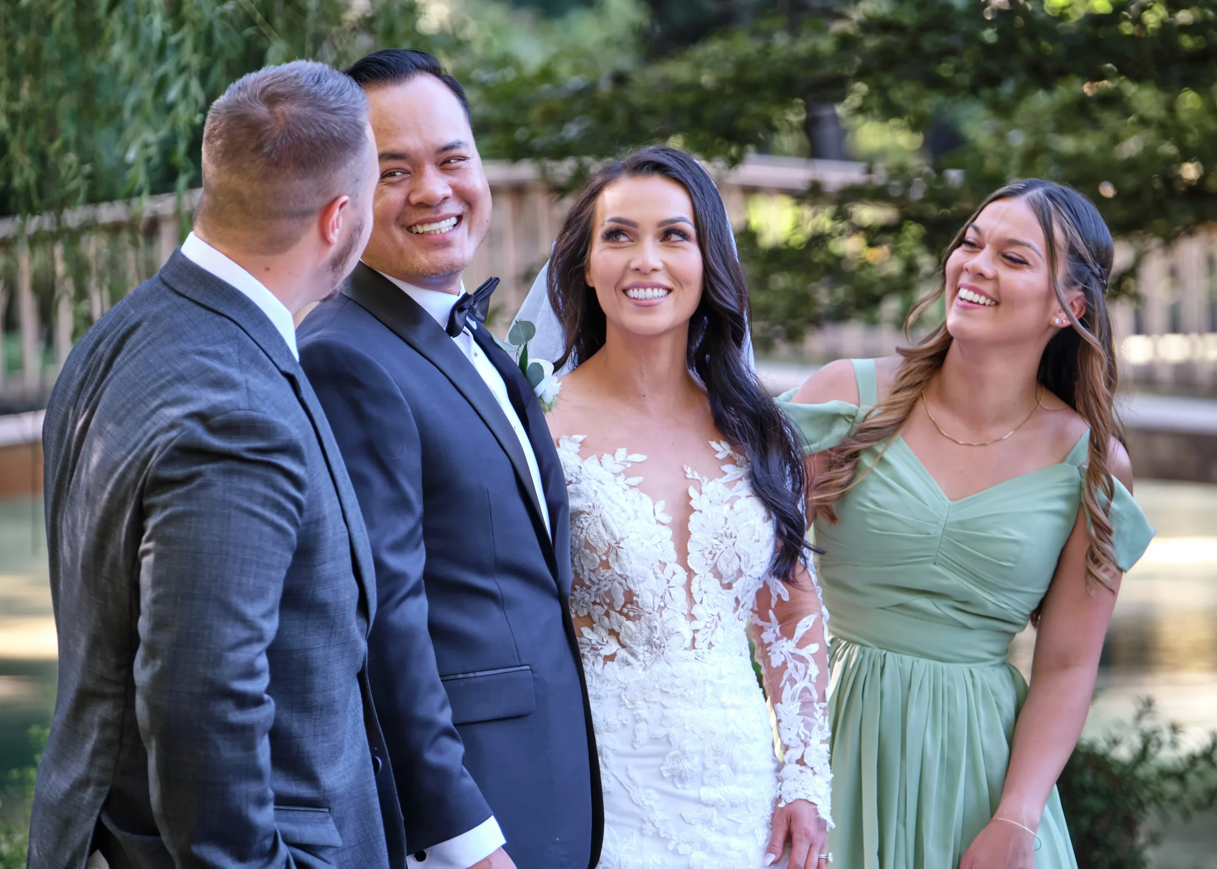 A wedding scene with four people outdoors. The bride and groom are in the center, smiling, surrounded by two women, all dressed in formal attire. There are trees and a fence in the background.