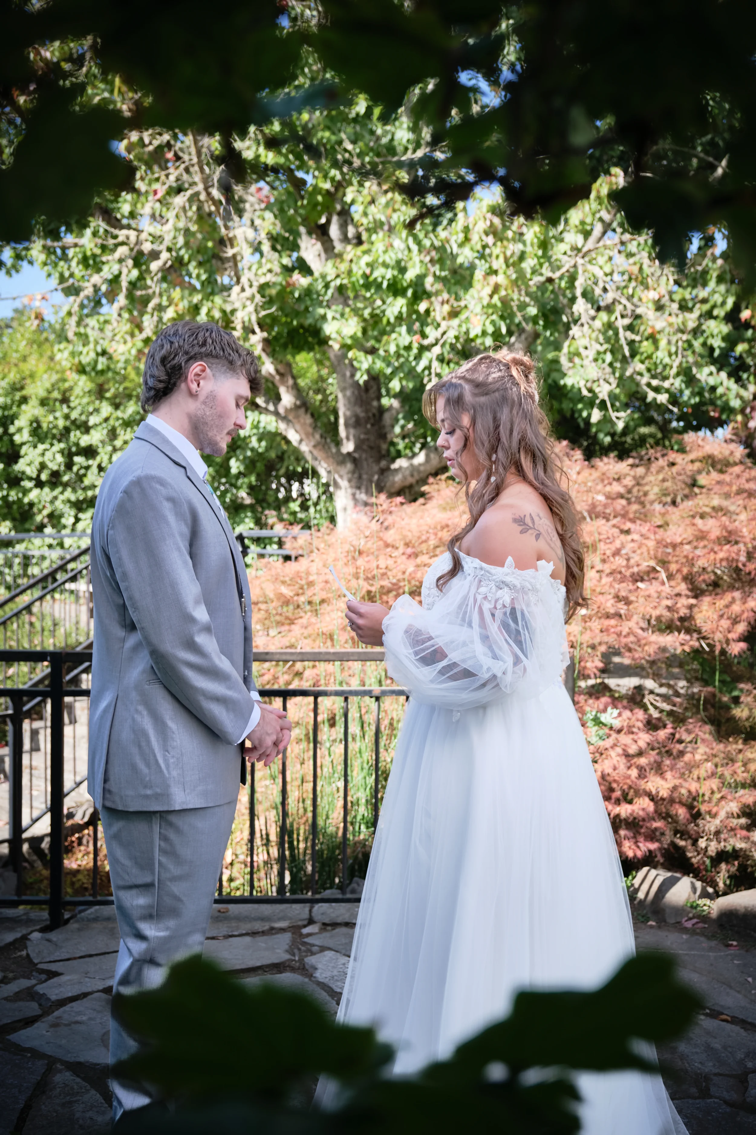 A man in a gray suit and a woman in a white wedding dress stand facing each other outdoors, exchanging vows in a garden setting with green trees and orange foliage.