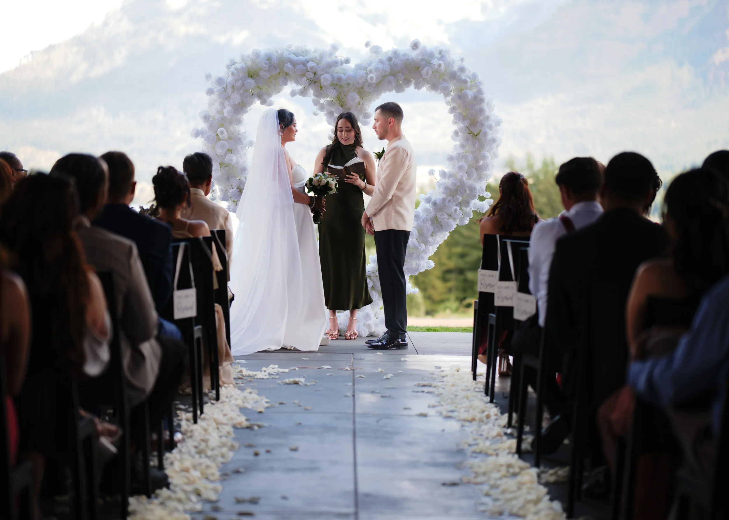 A wedding ceremony outdoors with the bride and groom holding hands, facing each other, under an arch decorated with white flowers. An officiant stands between them, reading from a book. Guests are seated on either side, watching the ceremony during d