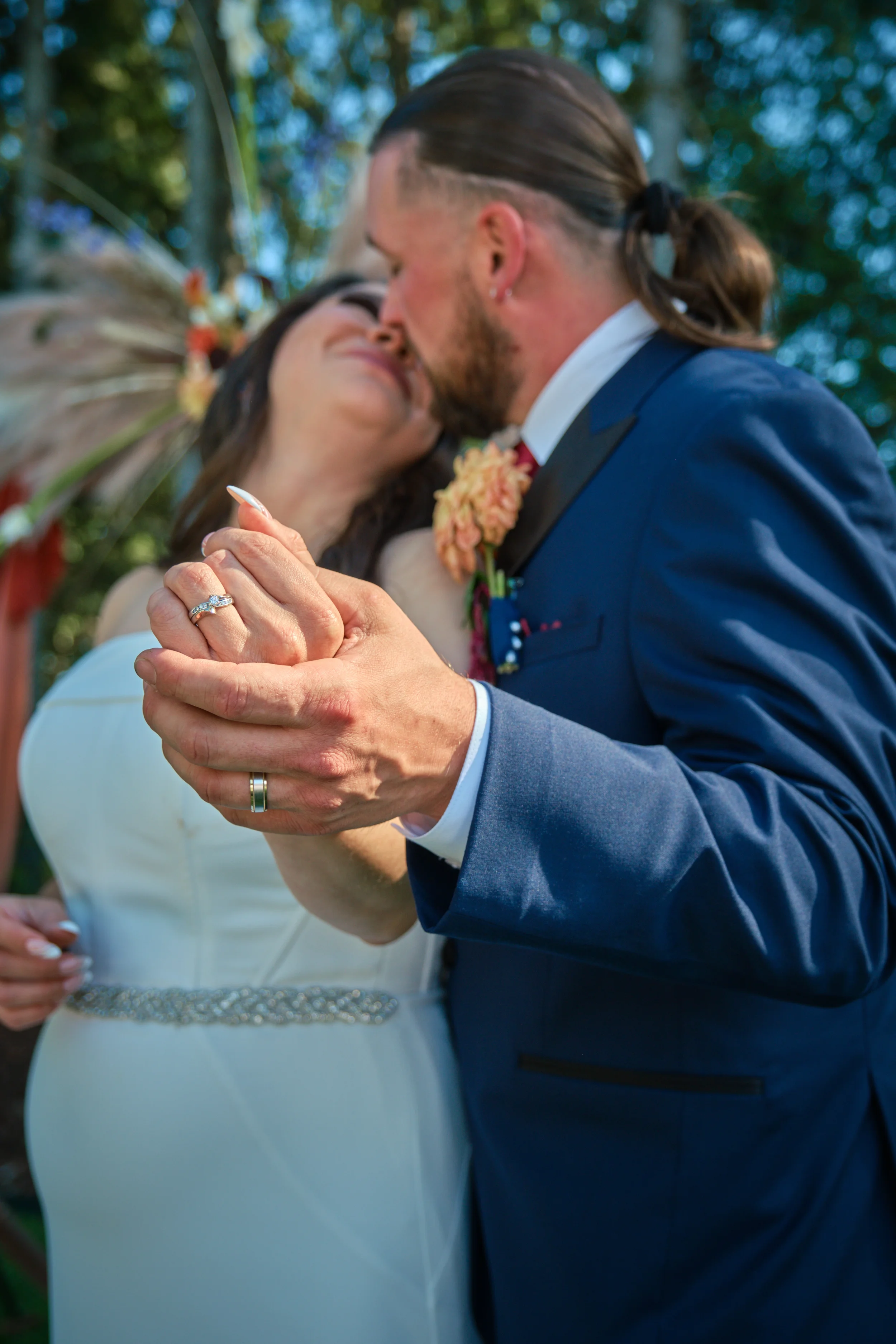 A couple is sharing a kiss at their outdoor wedding, holding hands, with a blurred background of trees.