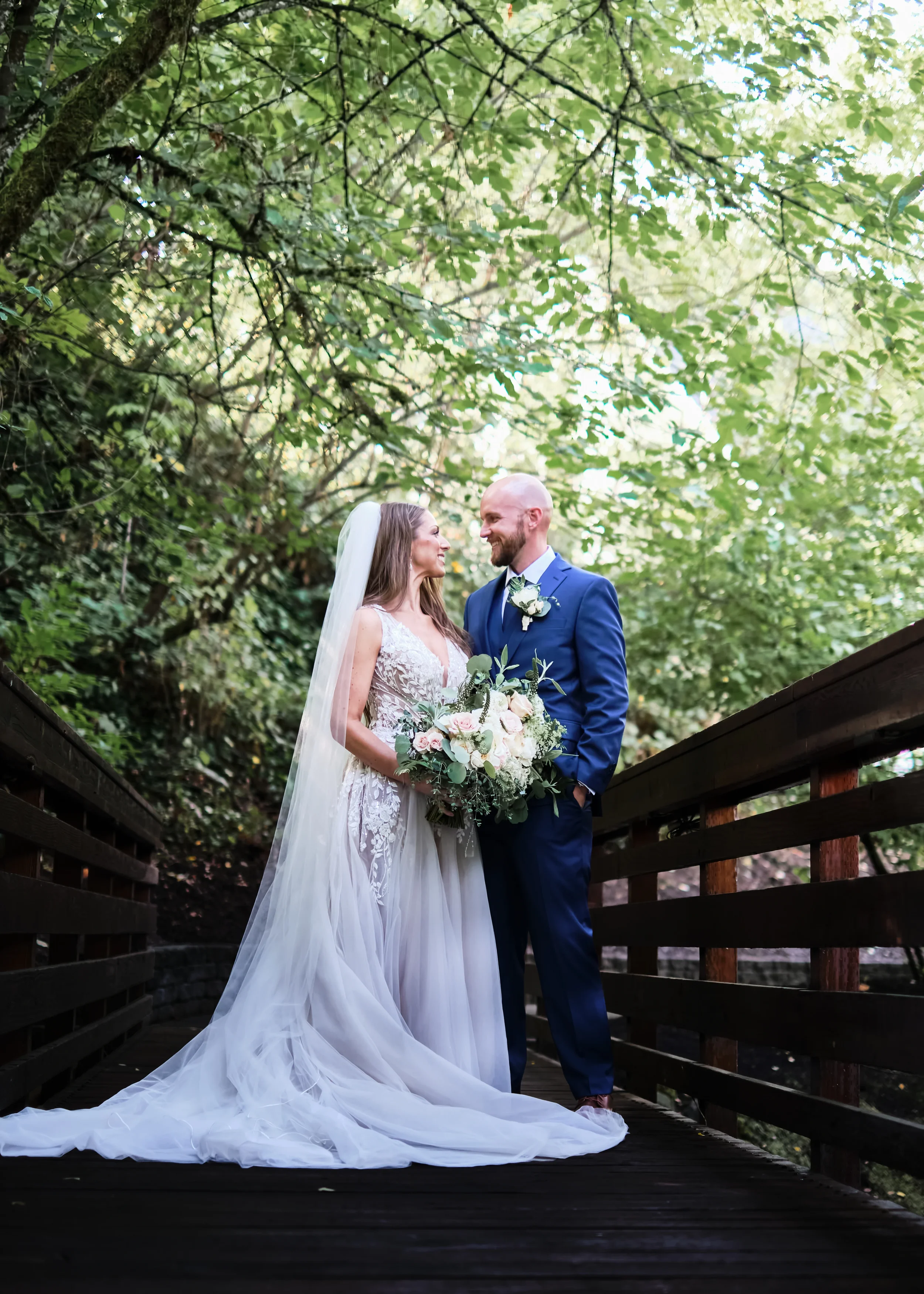 A bride and groom standing close together on a wooden bridge, smiling at each other in a lush green outdoor setting, with the bride holding a bouquet of flowers.