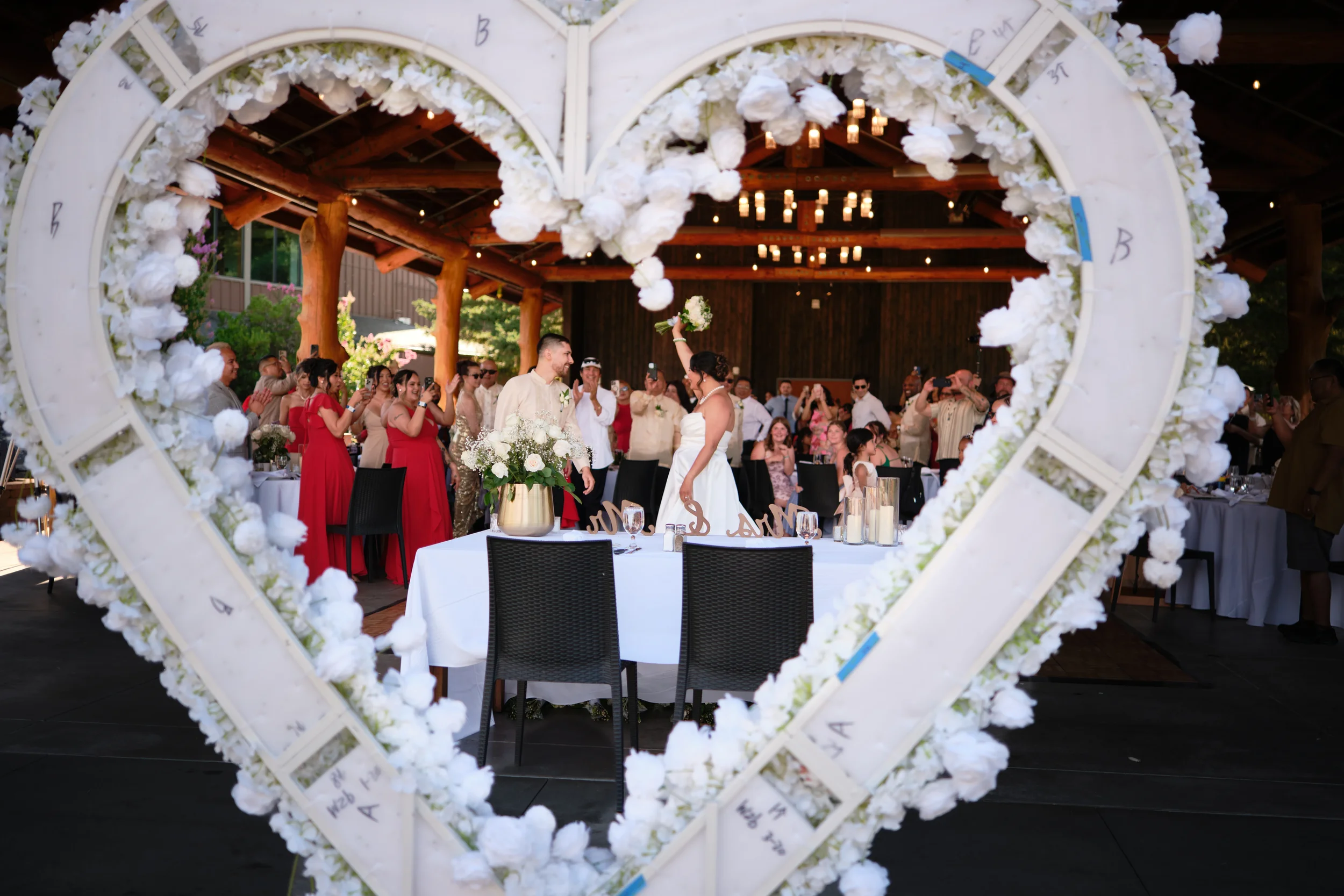 A wedding reception seen through a heart-shaped floral frame, with a bride and groom standing under a decorated ceiling, surrounded by guests.