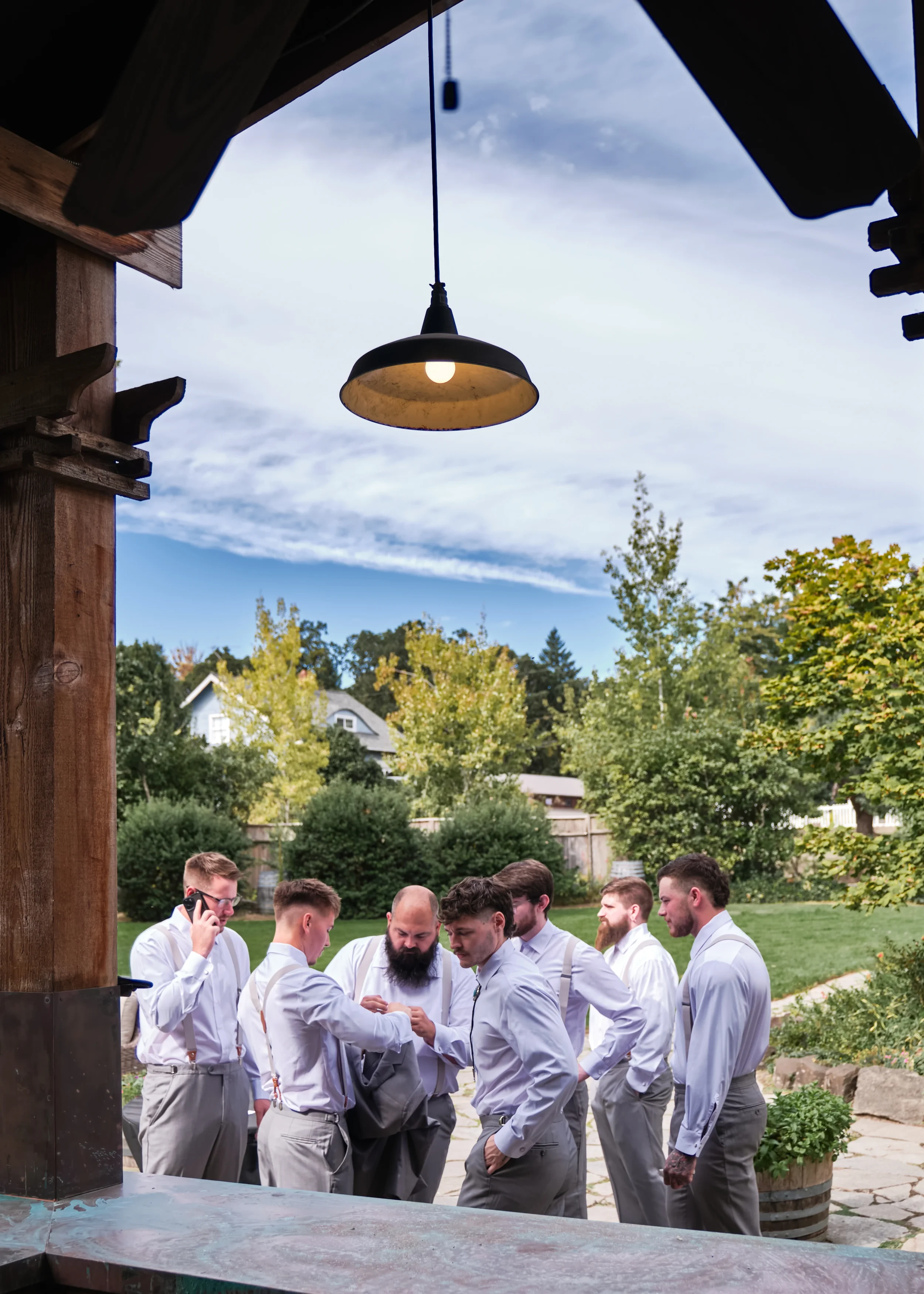 A group of men dressed in light gray suits and white shirts gathered outdoors in a garden or backyard setting, engaged in conversation or an activity, with trees, shrubs, and houses visible in the background under a partly cloudy sky.