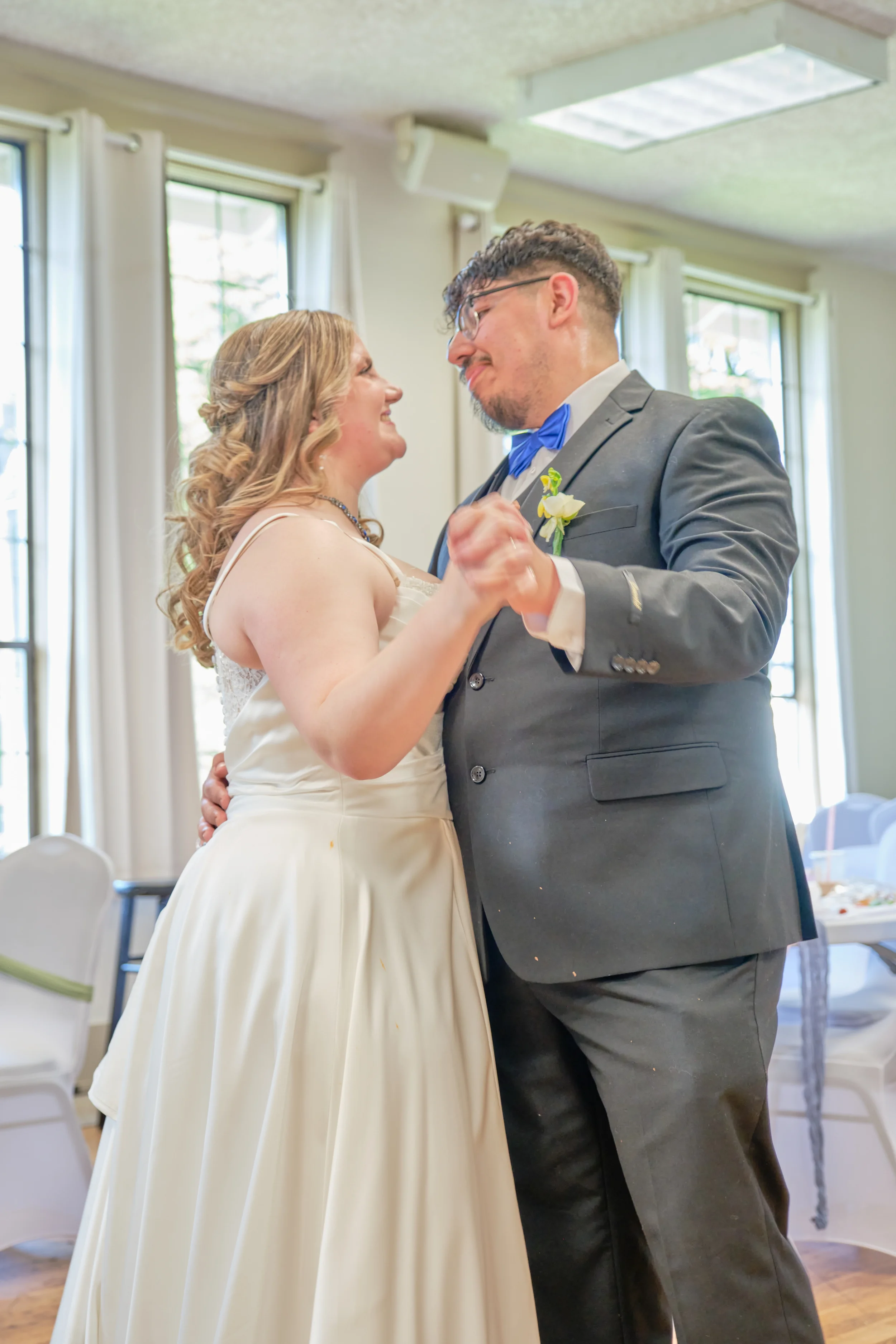 A bride and groom are dancing together during their wedding reception in a decorated room with white curtains and windows.