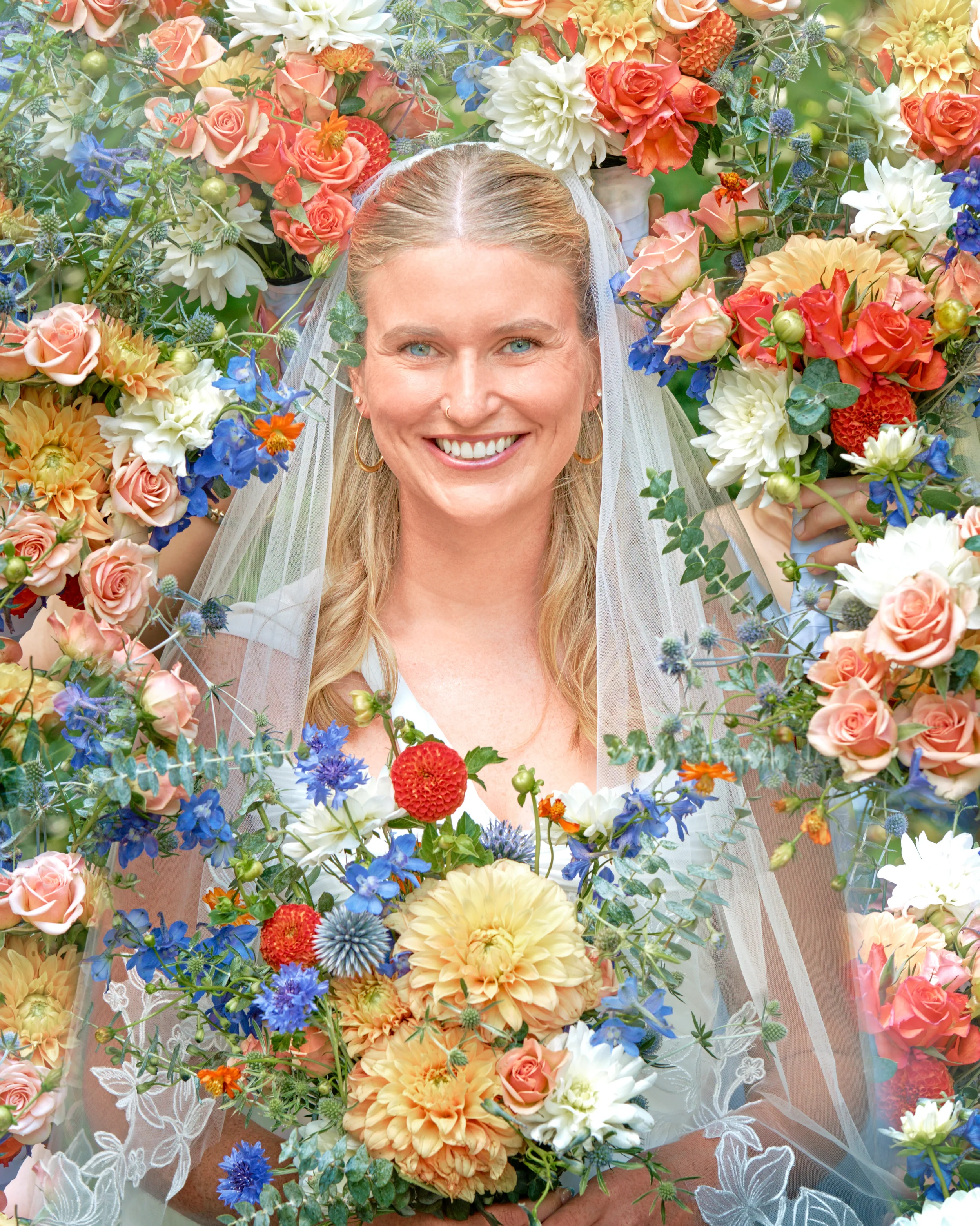 A woman smiling at the camera, wearing a veil, surrounded by a large arrangement of colorful flowers, including roses, dahlias, and other blooms in shades of pink, white, orange, blue, and purple.