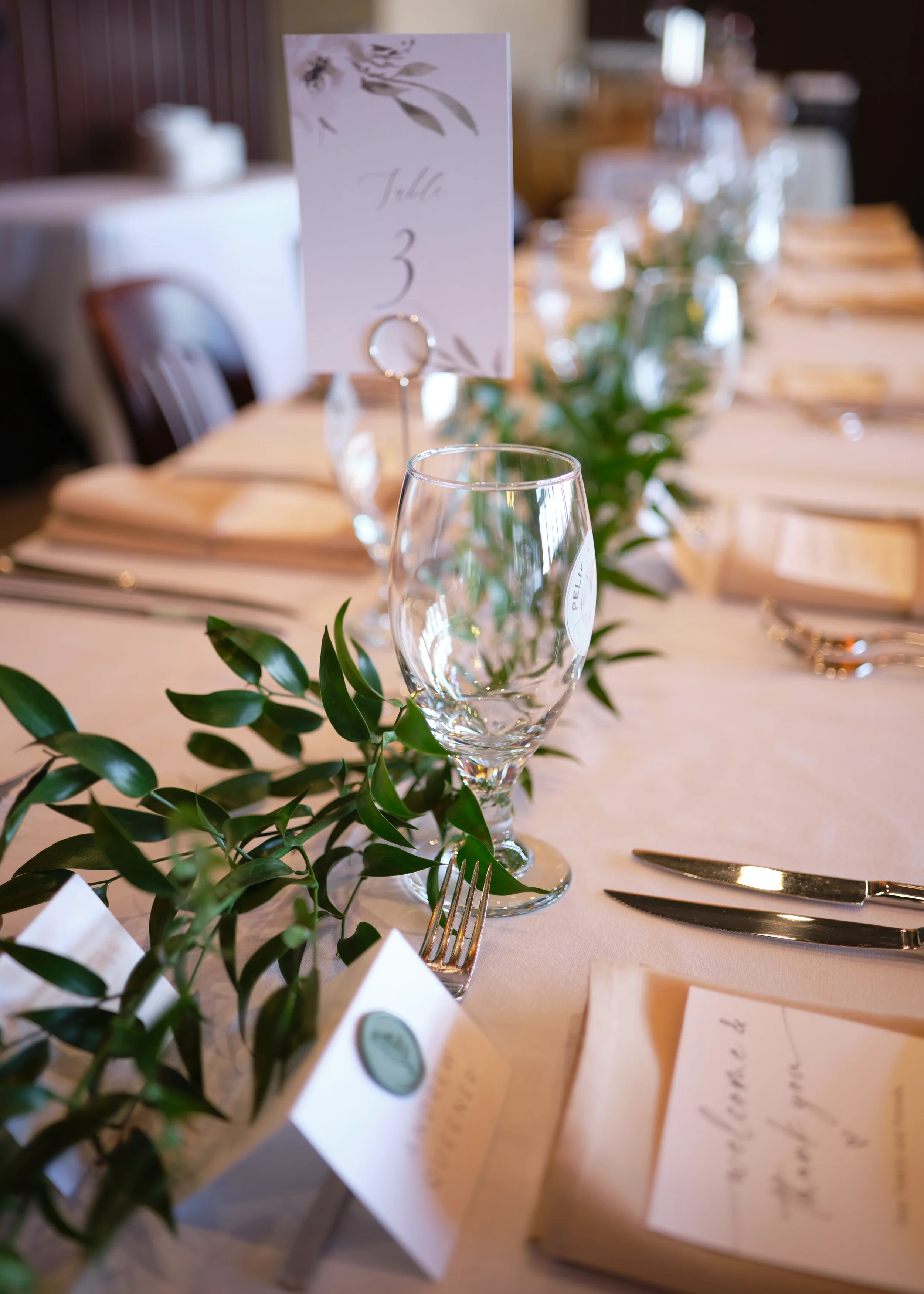 A close-up view of a decorated dining table with a wine glass, silverware, and a greenery garland. A small table number card with a floral design and handwritten text is visible in the background.