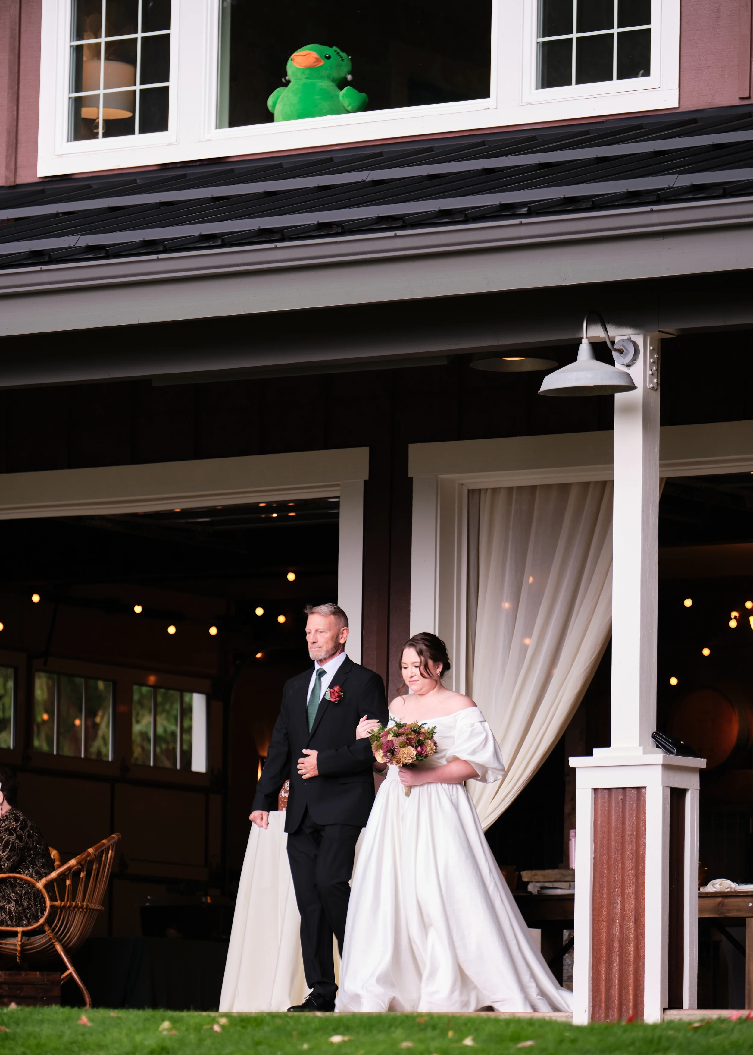 A bride in a white wedding dress holding a bouquet of flowers is walking with an older man in a dark suit, likely her father, at a wedding reception held in a rustic venue. A humorous green duck plush toy is seen at the window of the building.