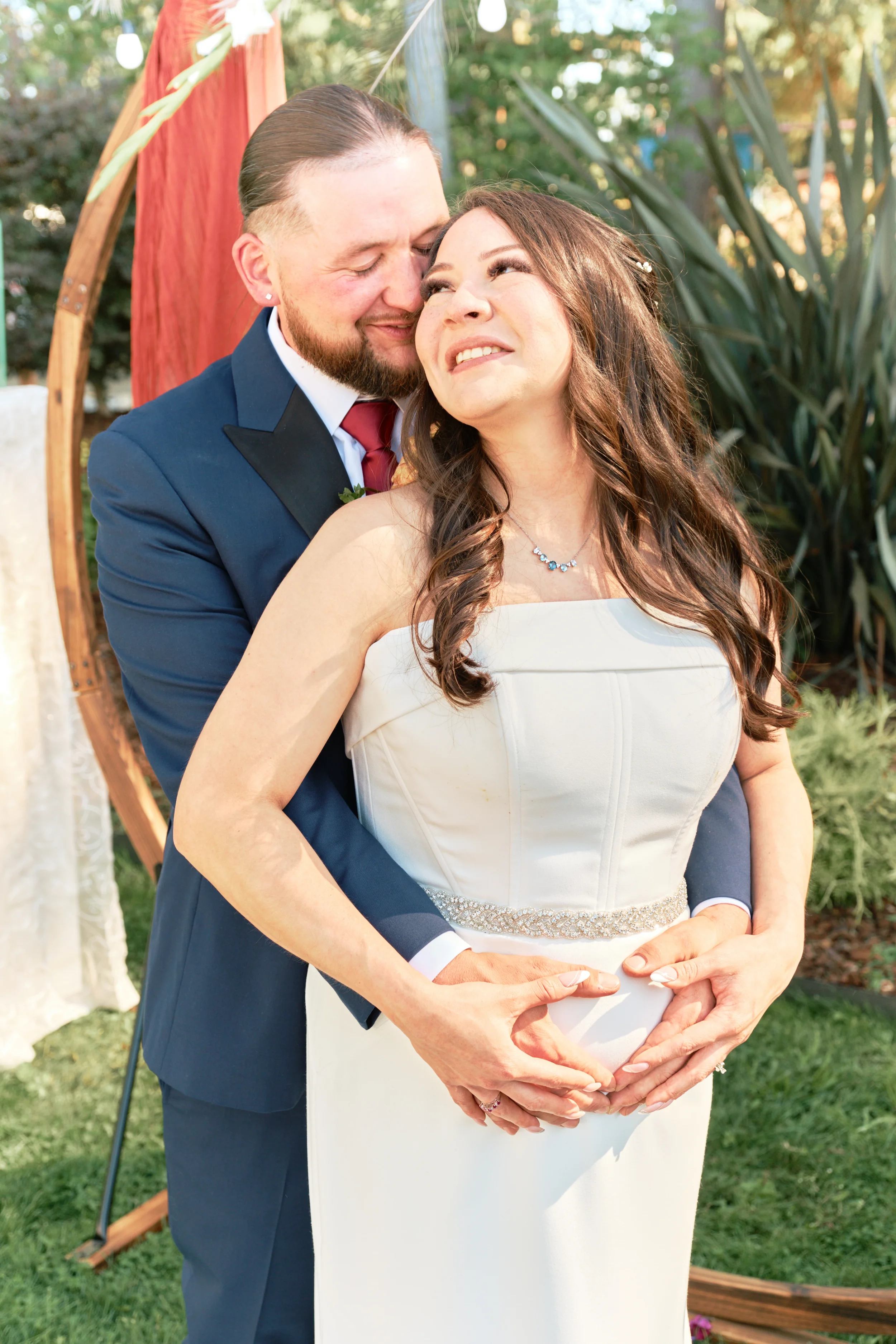 A newlywed couple at their outdoor wedding, with the groom in a navy suit and the bride in a white strapless dress, smiling and holding her baby bump.