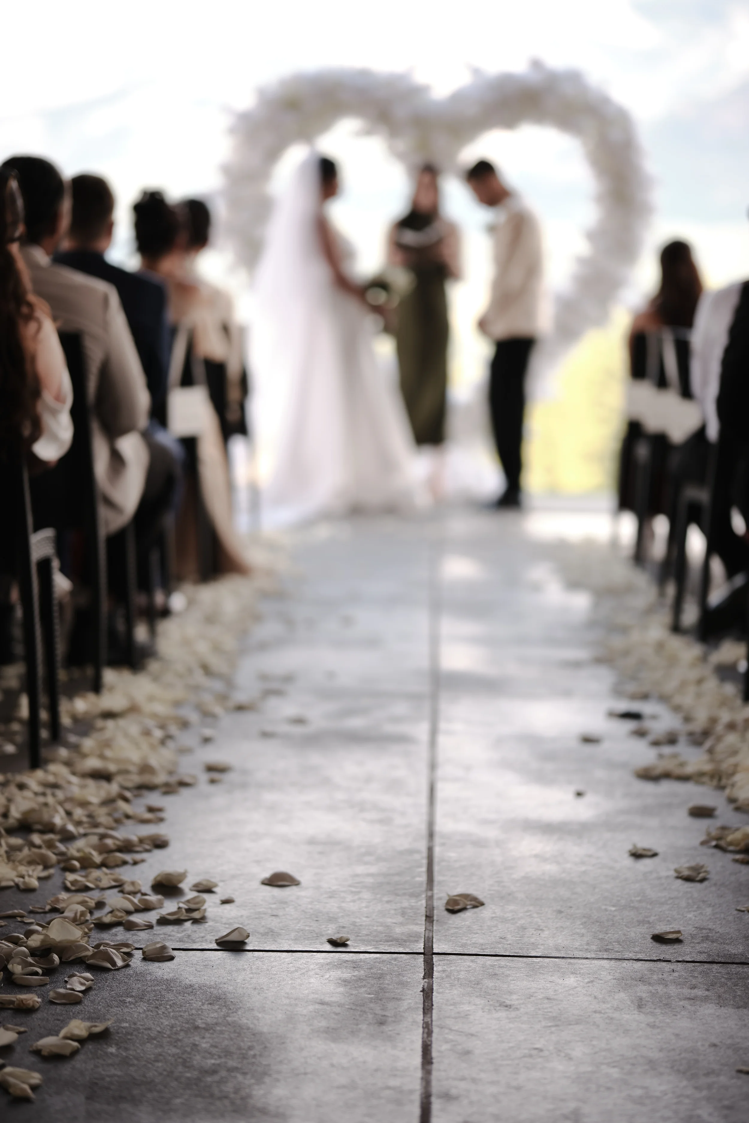A wedding ceremony with a couple holding hands in front of an officiant, under a heart-shaped floral arch, with guests seated on either side, and petal scatter on the aisle.