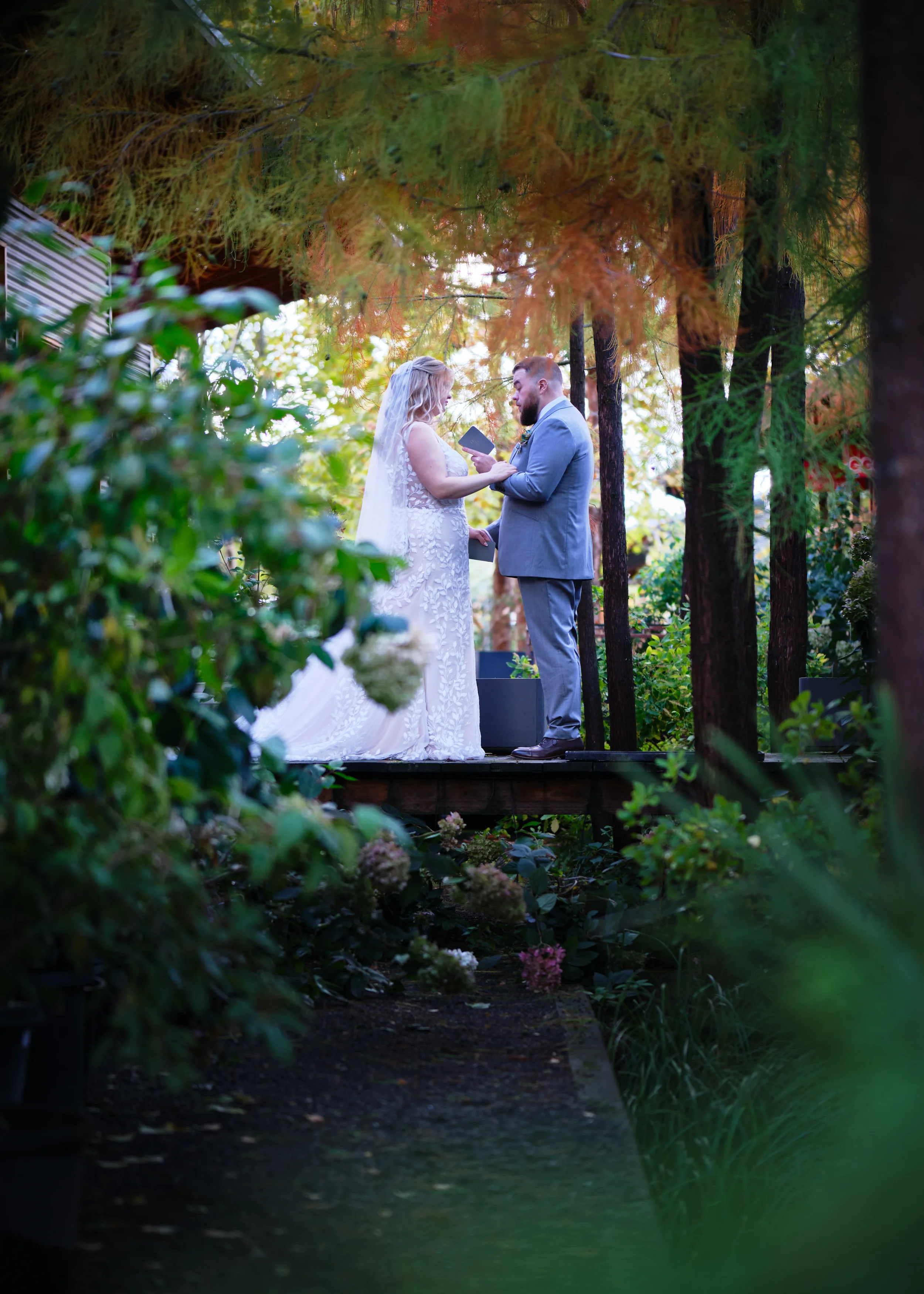 A couple exchanging vows or rings during a wedding ceremony in an outdoor forest setting surrounded by tall trees and lush greenery.