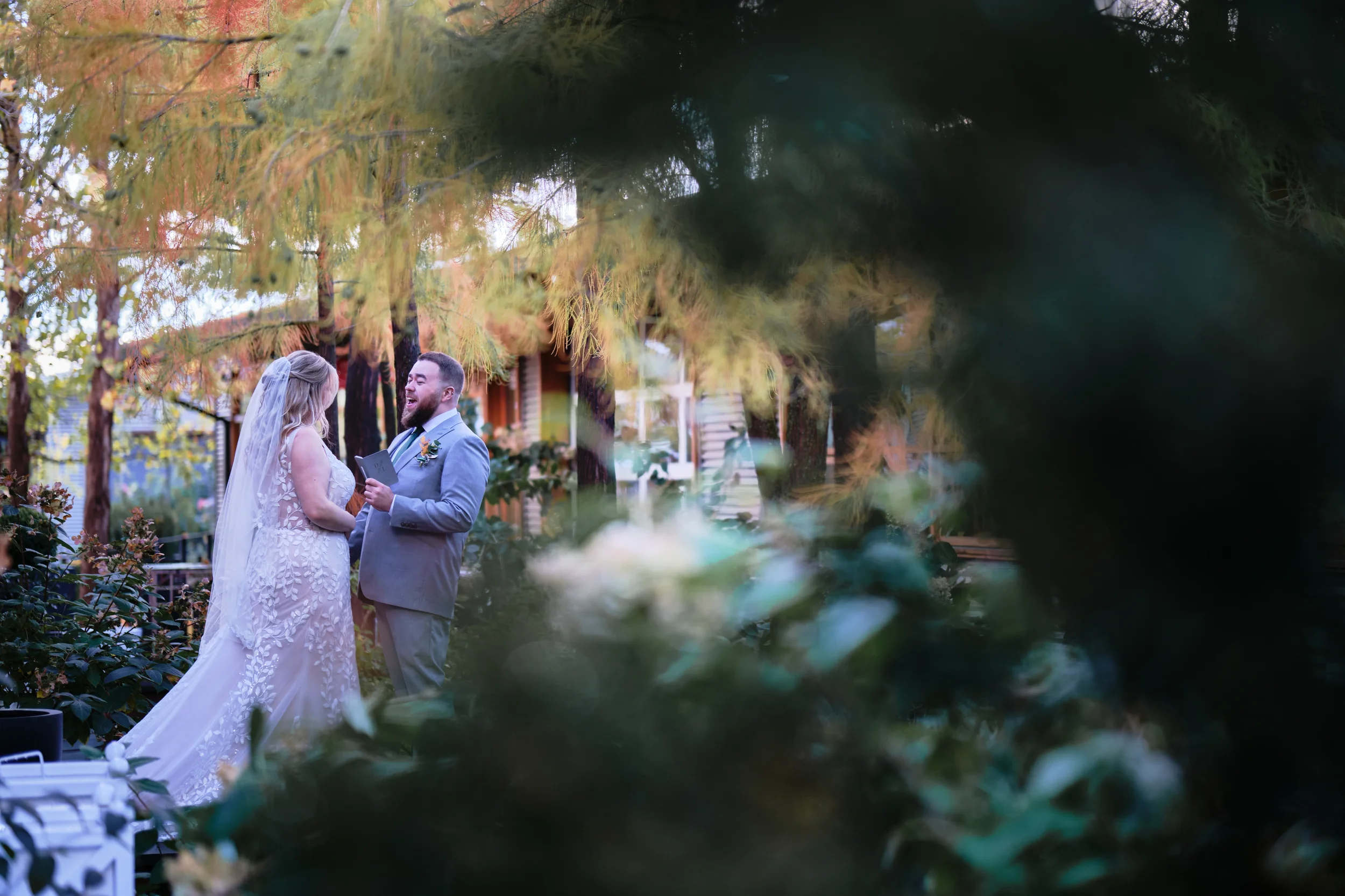 A bride and groom standing outdoors during their wedding ceremony, surrounded by trees and greenery, sharing a joyful moment.