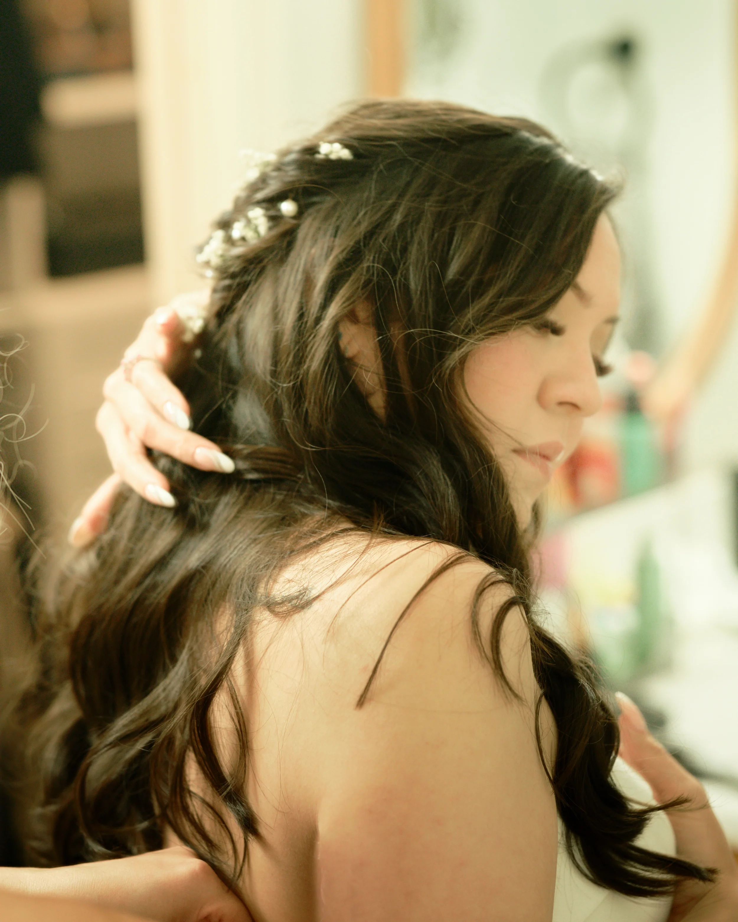 Close-up of a woman with dark, wavy hair decorated with small white flowers, holding her hair with one hand, with a soft background.