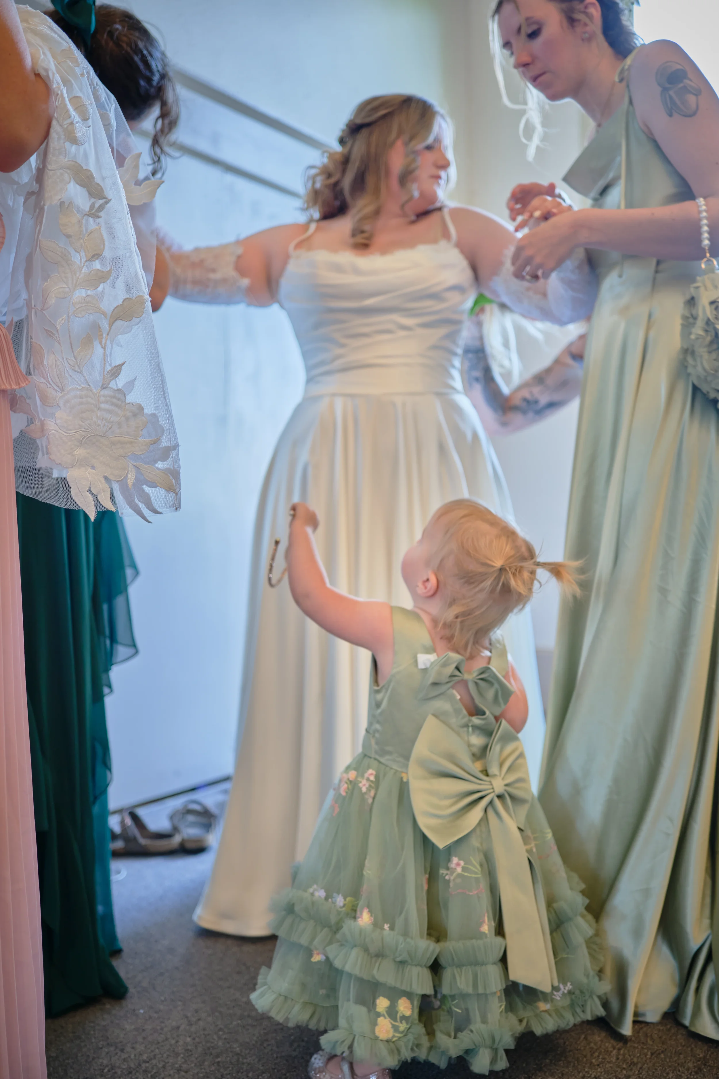 A woman in a white wedding dress is being helped by another woman in a light green dress, with a young girl in a green dress wearing a large bow, in a room with a carpeted floor and shoes near the door.
