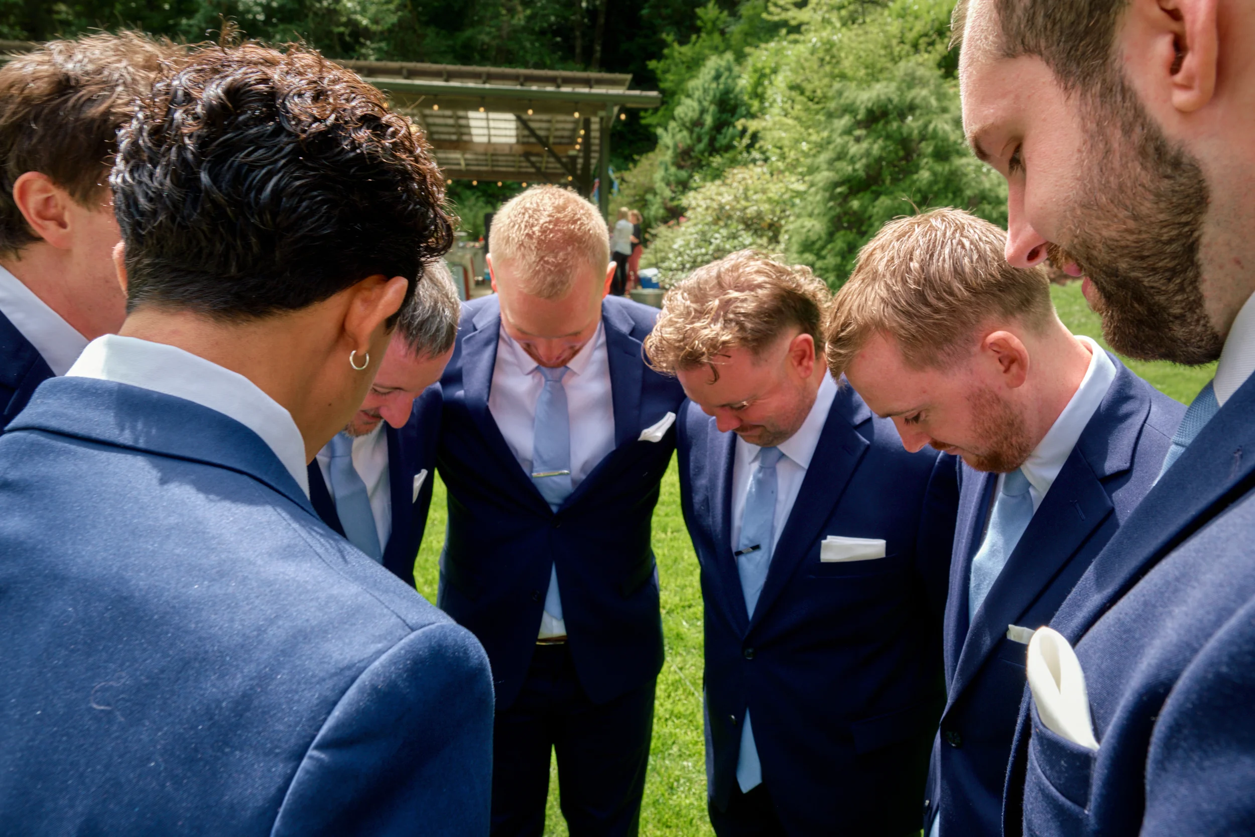 Group of men dressed in suits, standing in a circle outdoors with heads bowed in prayer or reflection.