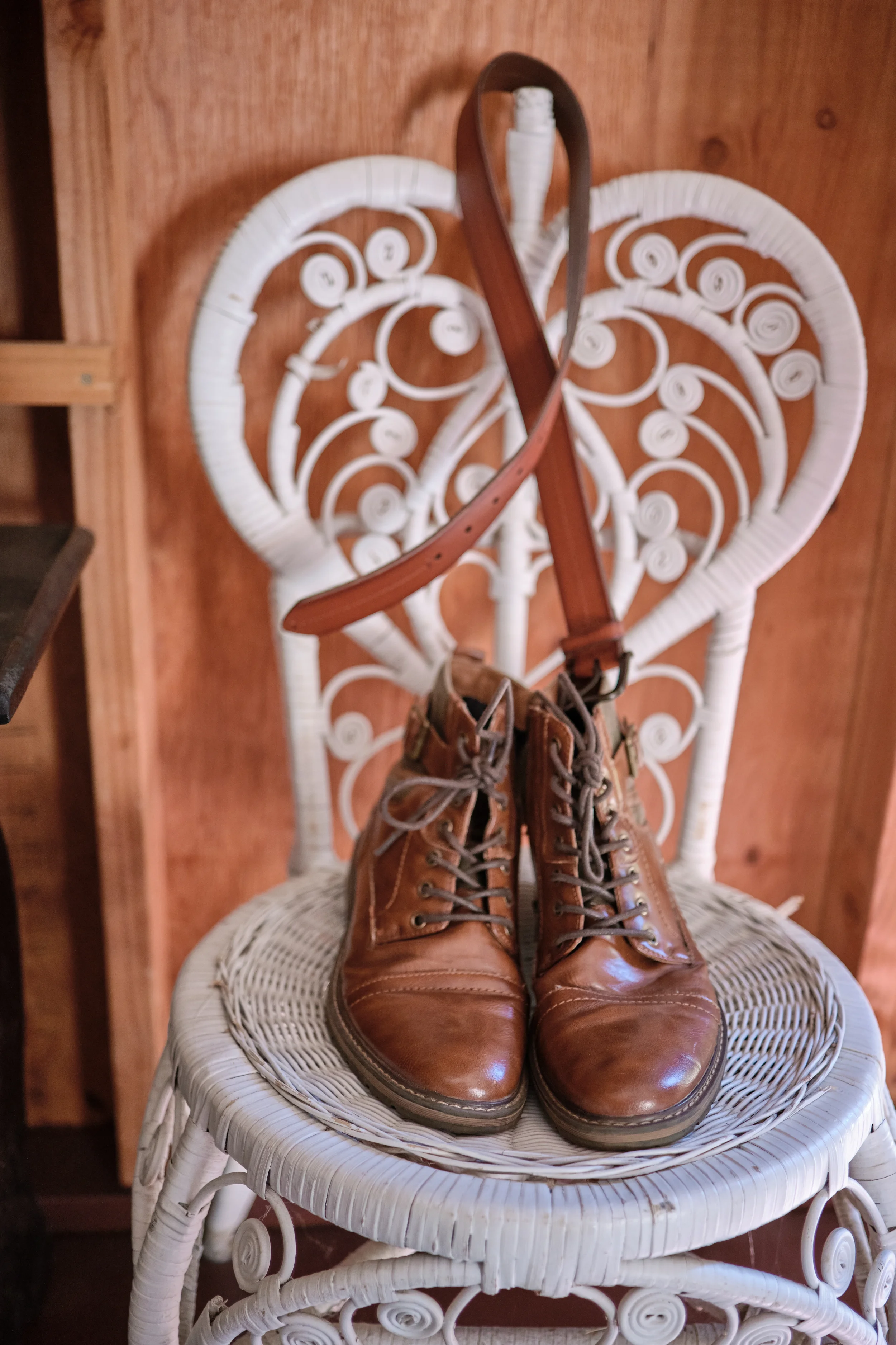 A pair of brown leather lace-up boots placed on a white ornate chair with a wooden backrest, in front of a wooden wall.