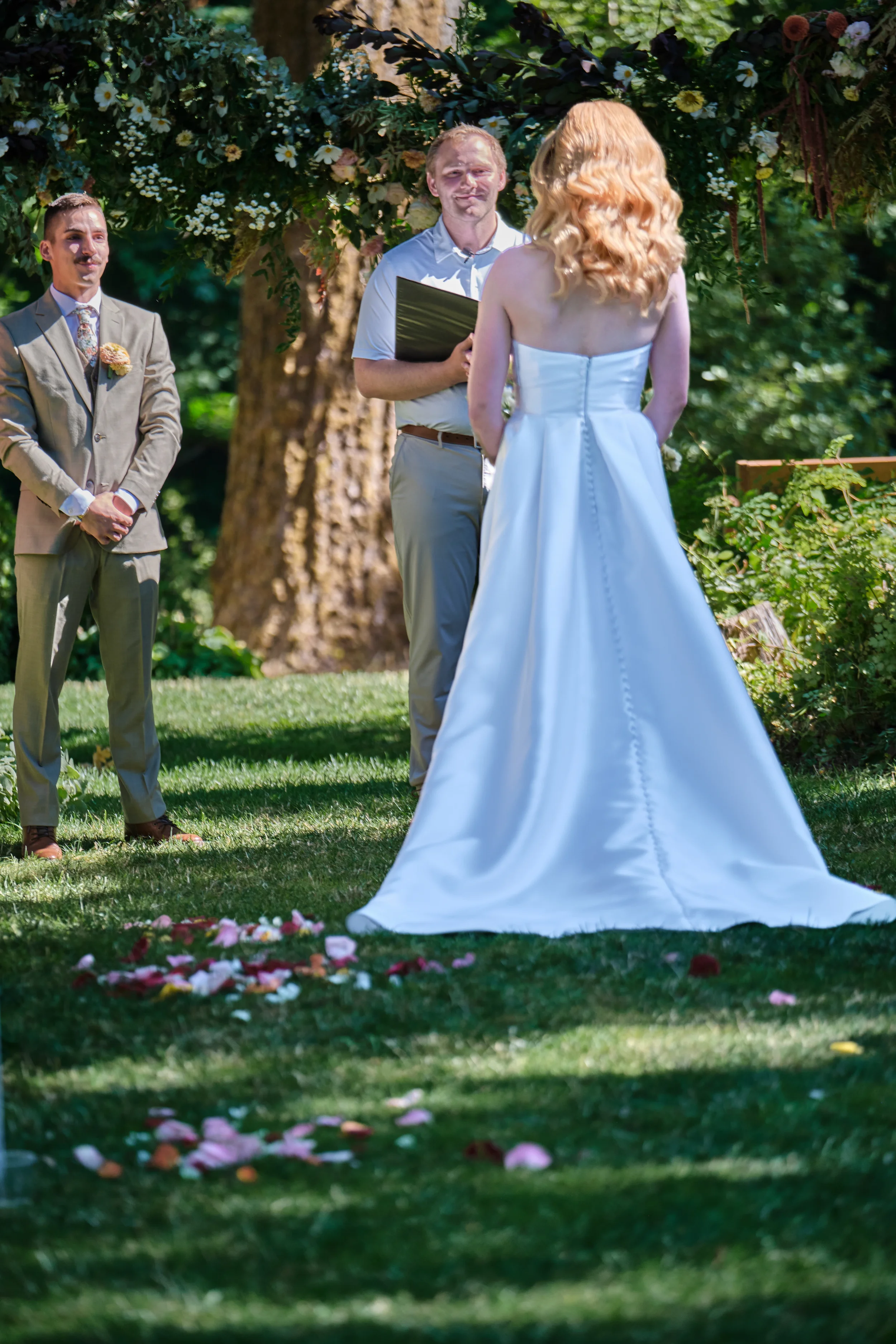 A wedding ceremony taking place outdoors under a large tree decorated with flowers, with a bride and groom facing each other and an officiant holding a folder.