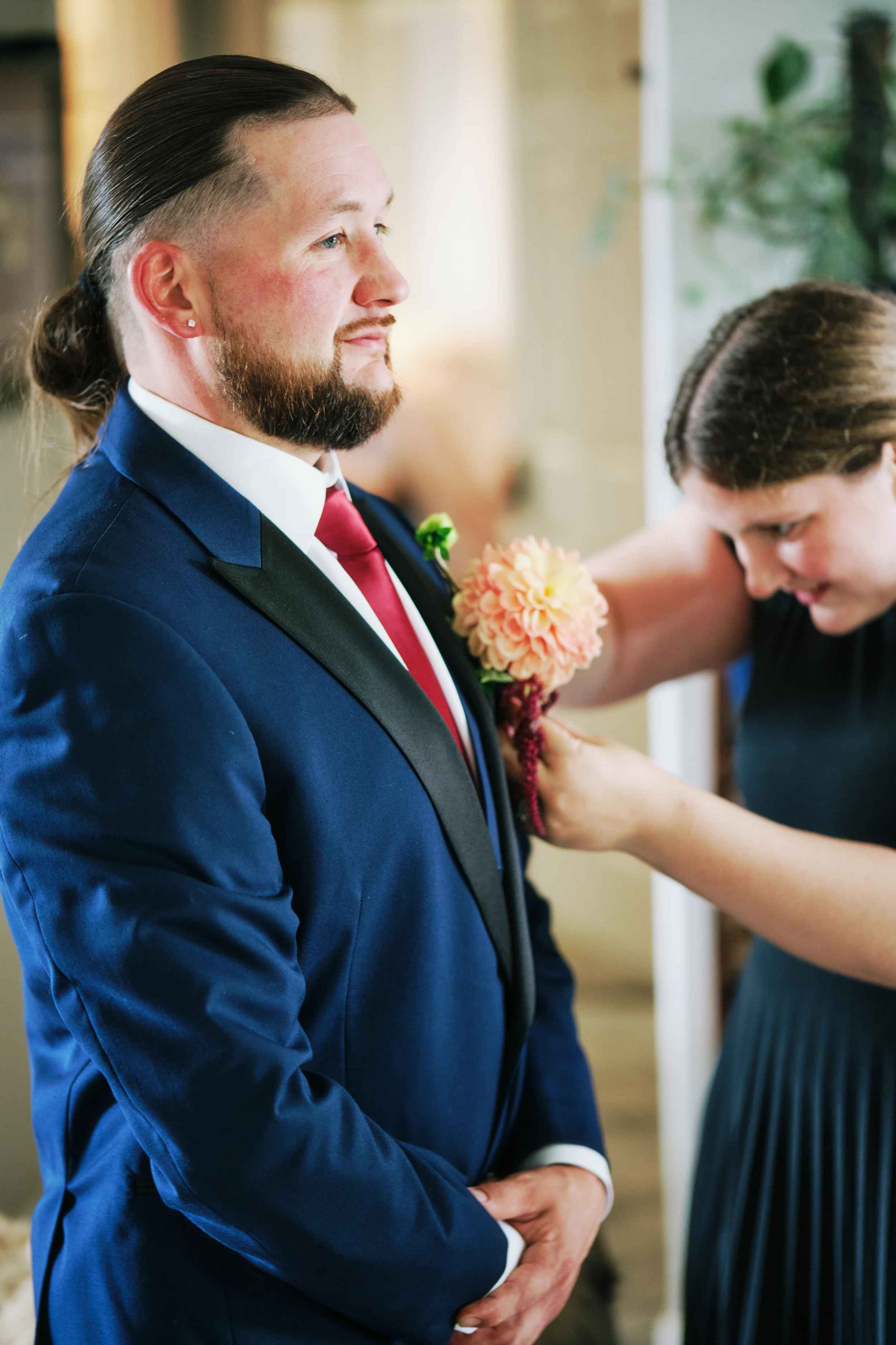 Man wearing a blue suit with a red tie and boutonniere receiving a hug from a woman, during a wedding or special event.
