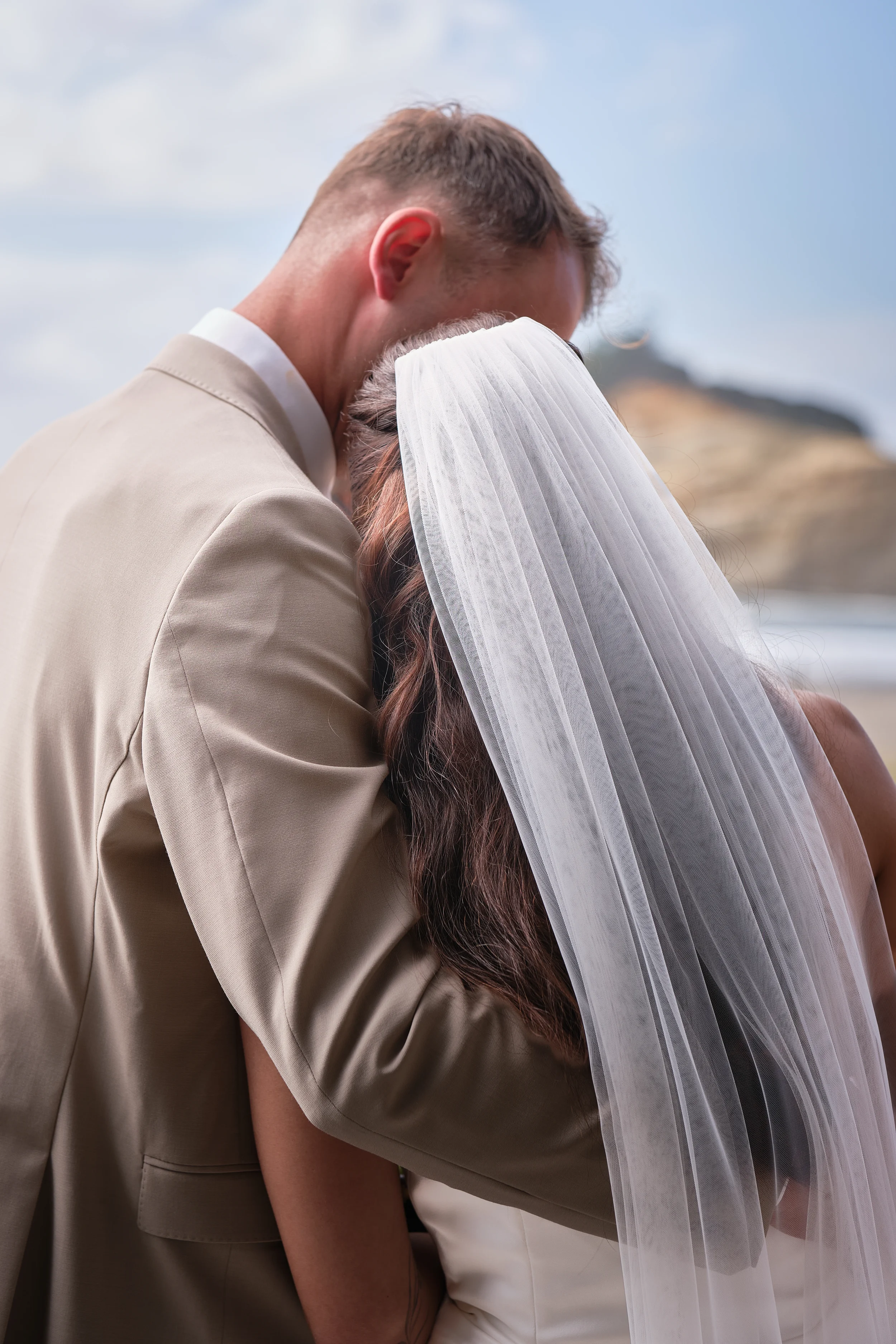 A couple, the man wearing a beige suit and the woman in a white dress with a veil, are embracing outdoors with a beach and rocky hill in the background.