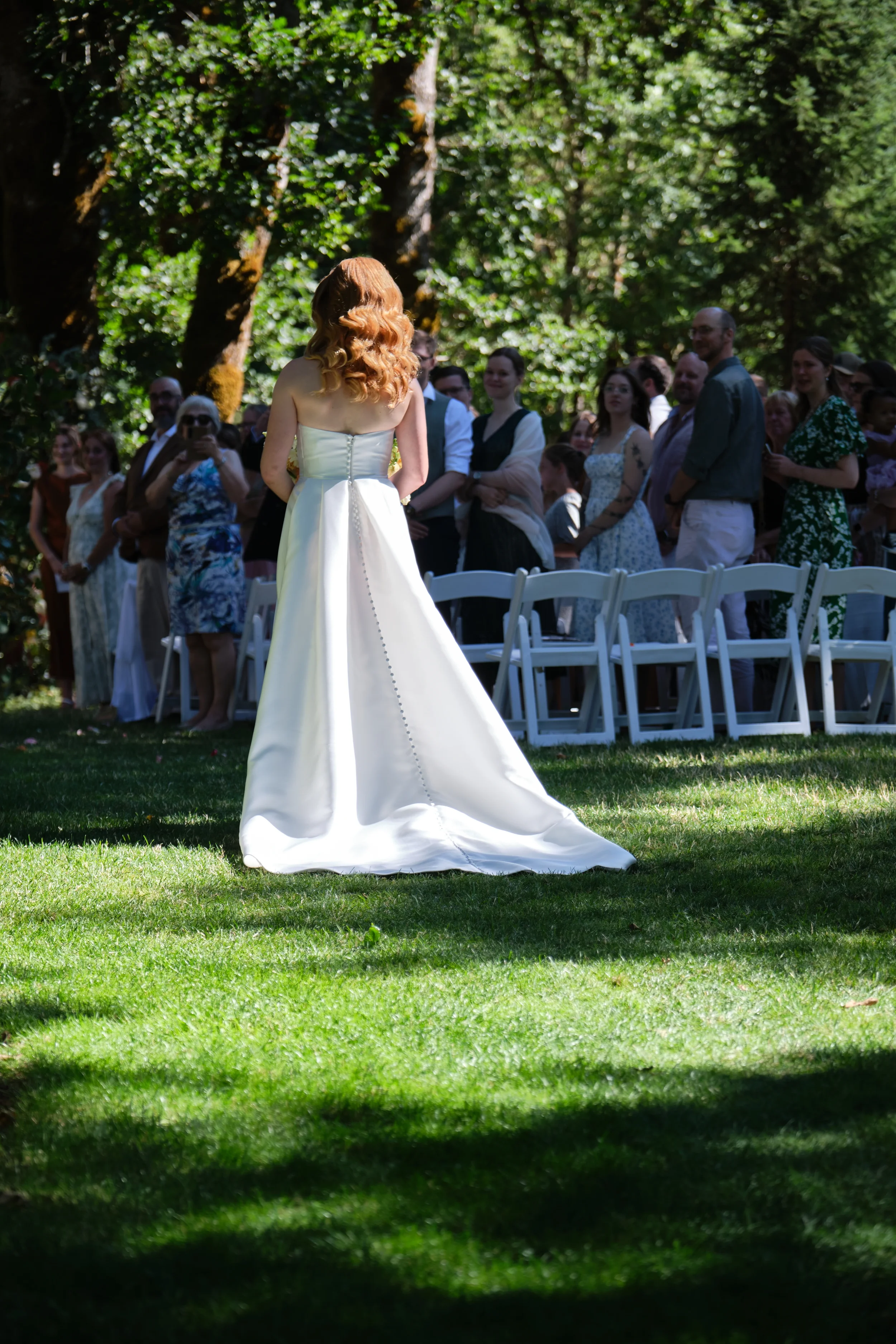 A bride in a white wedding gown with red hair stands outdoors on a grassy area, facing a group of guests. The guests are standing behind white chairs, watching her. The background features large trees and lush green foliage, with sunlight filtering t