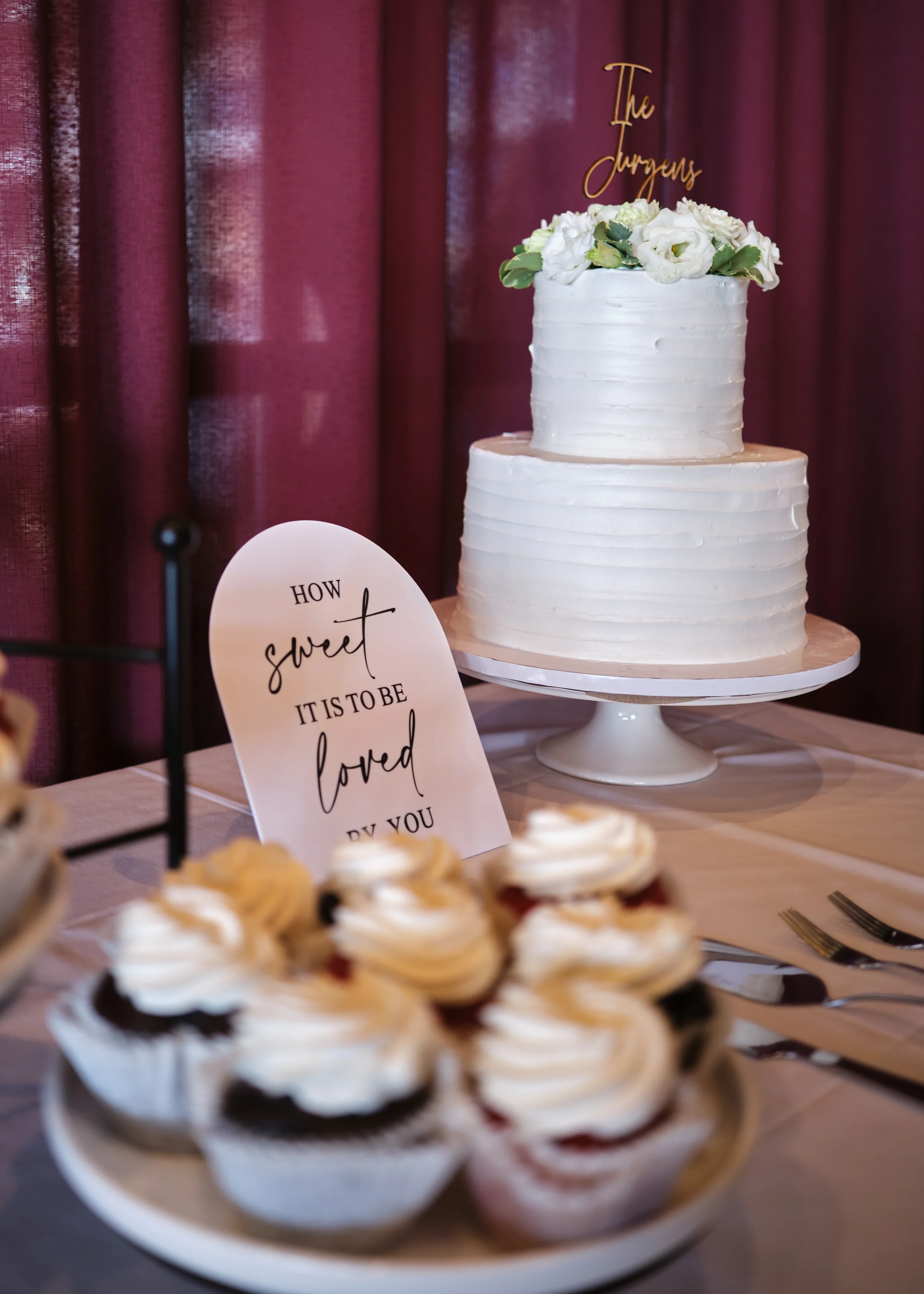 Two-tiered white wedding cake decorated with white flowers and greenery, with a gold topper reading 'The Jurgens' in front of maroon curtains.