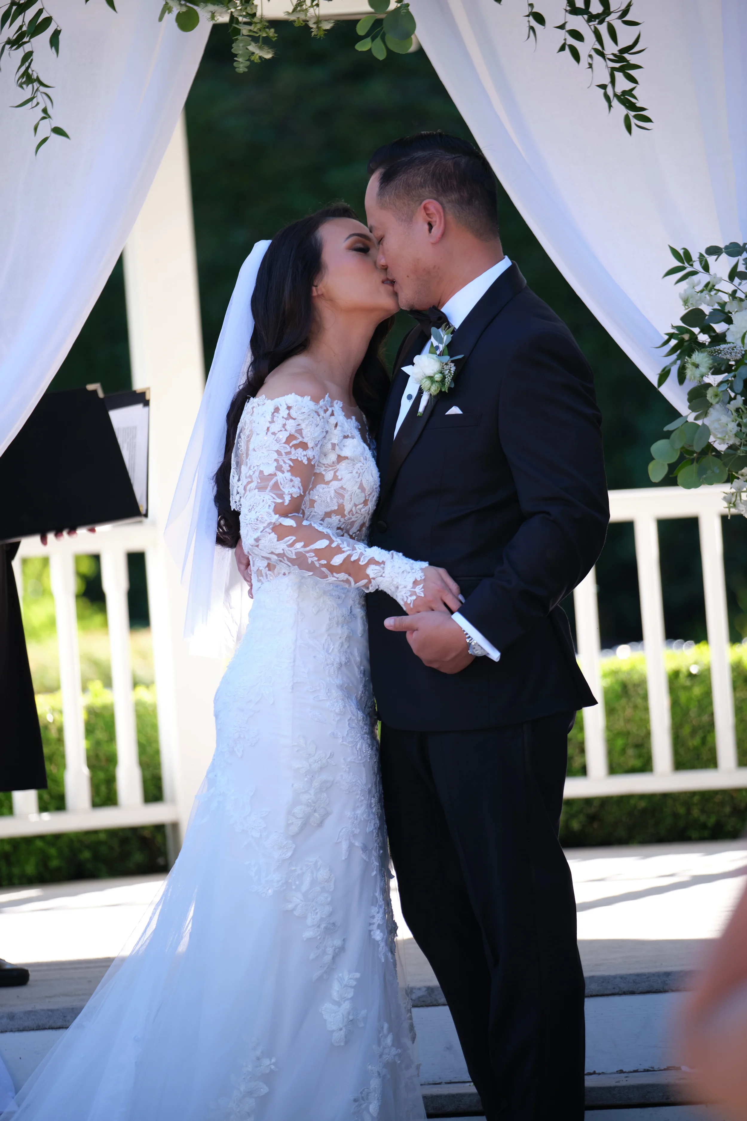 A bride and groom sharing a kiss under a wedding arch decorated with greenery and white flowers.