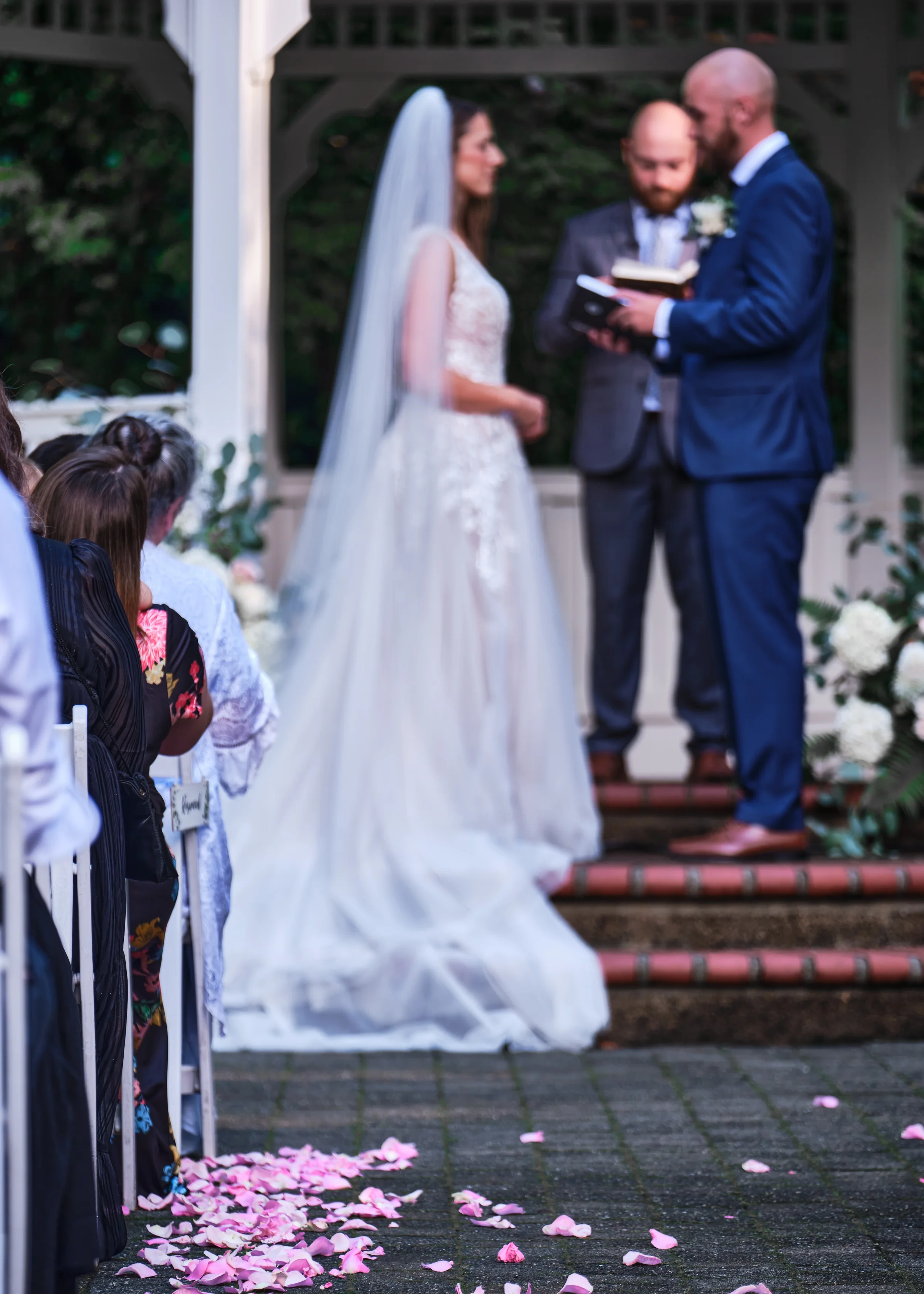 A wedding ceremony taking place outdoors, with pink flower petals scattered on the ground. The bride and groom are standing before an officiant, who is reading from a book. Guests are seated on the side, watching the ceremony.