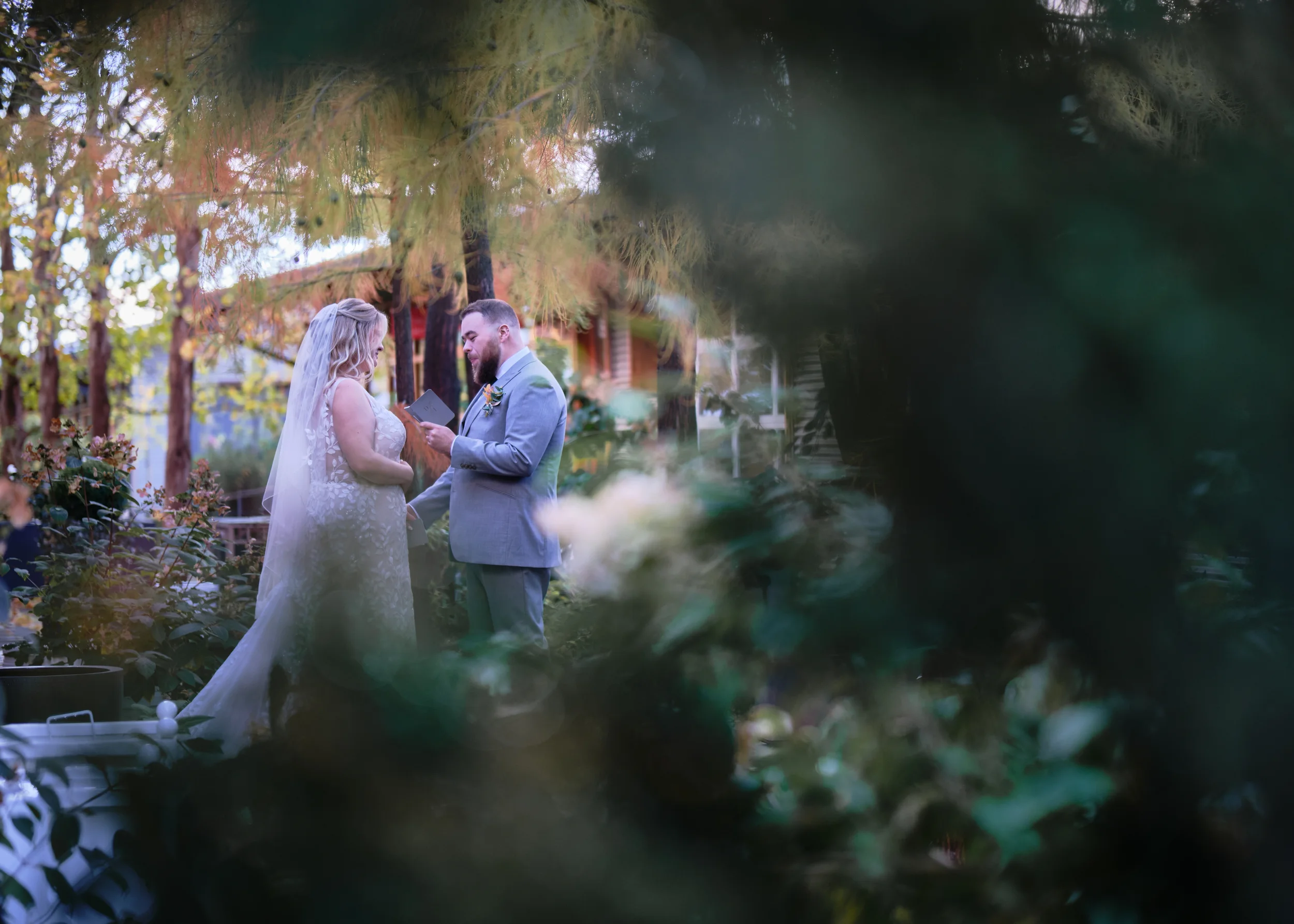 A bride and groom exchanging vows during a wedding ceremony outdoors surrounded by trees and greenery, seen through a blurred foreground.