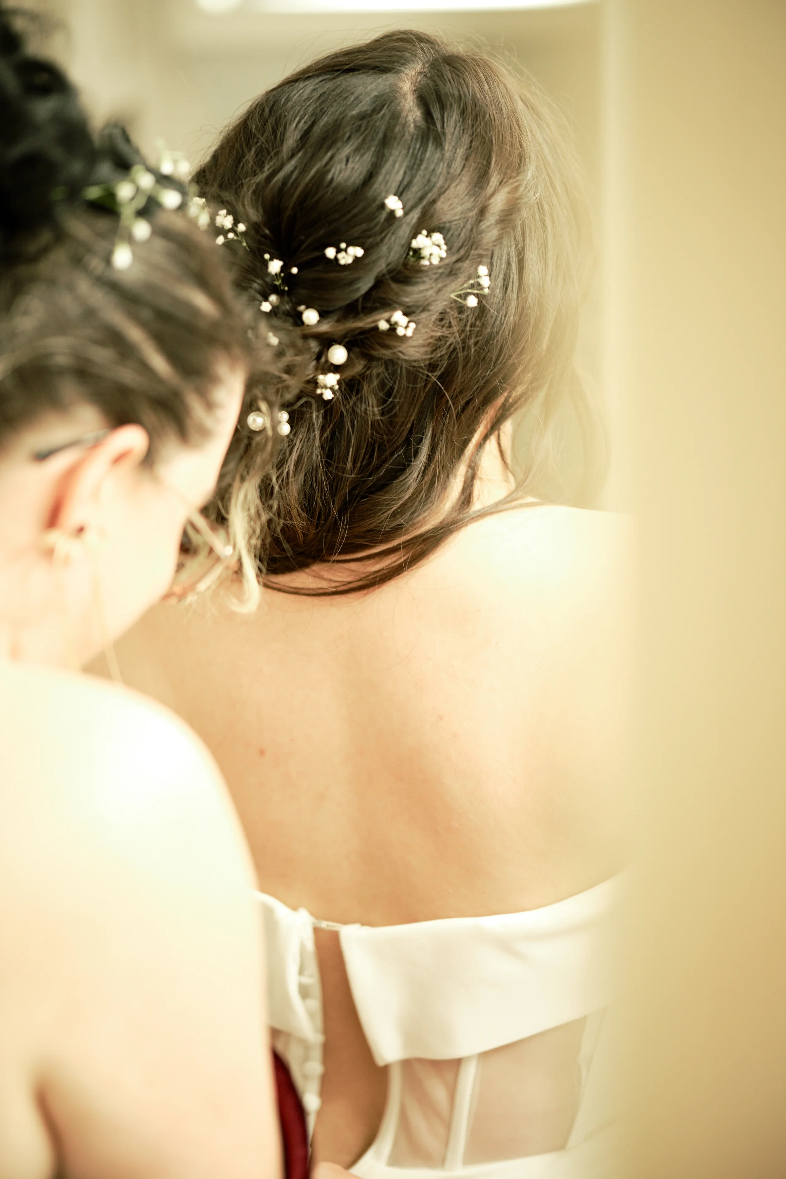 Close-up of a woman's back with brown hair decorated with small white flowers, wearing a white strapless top, and partially visible person with dark hair in the foreground.