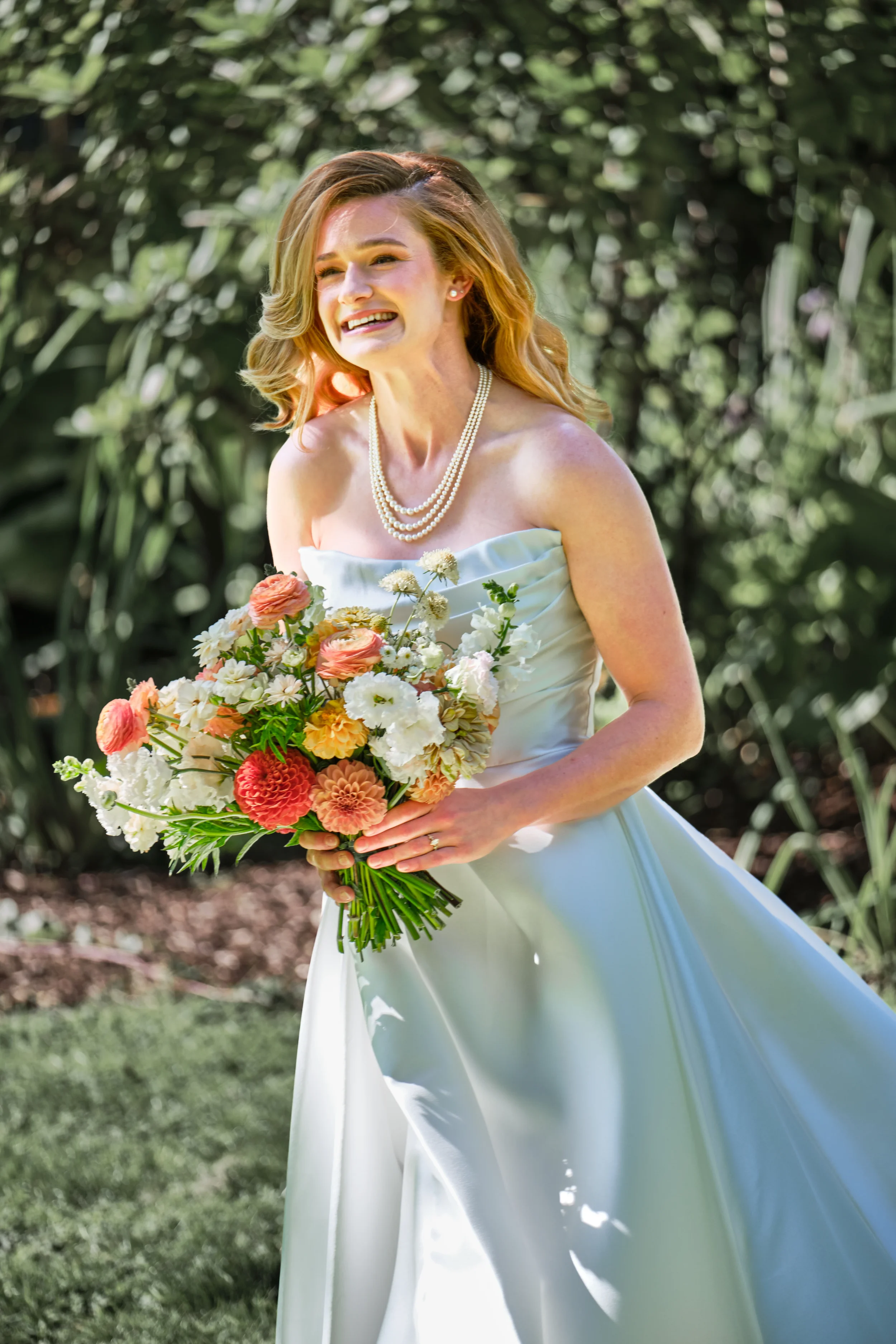 A woman in a white strapless dress holding a bouquet of colorful flowers outdoors, smiling and enjoying her time.