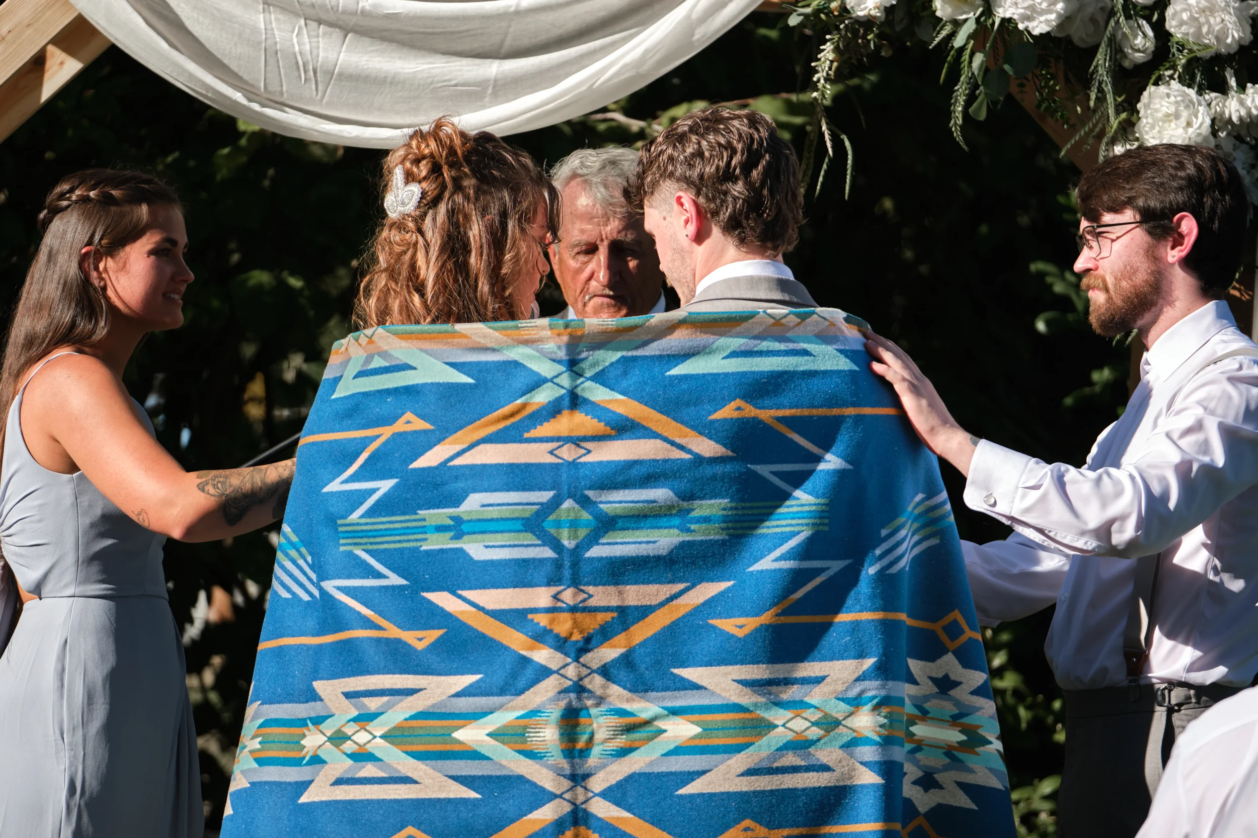 A group of six people participating in a wedding ceremony outdoors surrounded by greenery. Four participants are facing each other, with two women on the left and two men on the right. The bride has curly red hair and is wearing a hair accessory, and