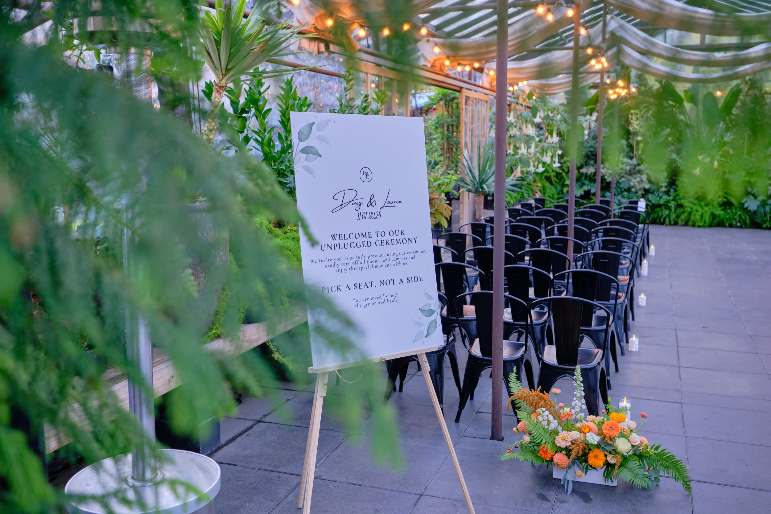 Wedding ceremony setup outdoors with chairs arranged in rows, floral arrangements, and a welcome sign that reads 'Doug & Lauren, 11.01.2025, Welcome to our Unplugged Ceremony' in a garden setting with string lights.