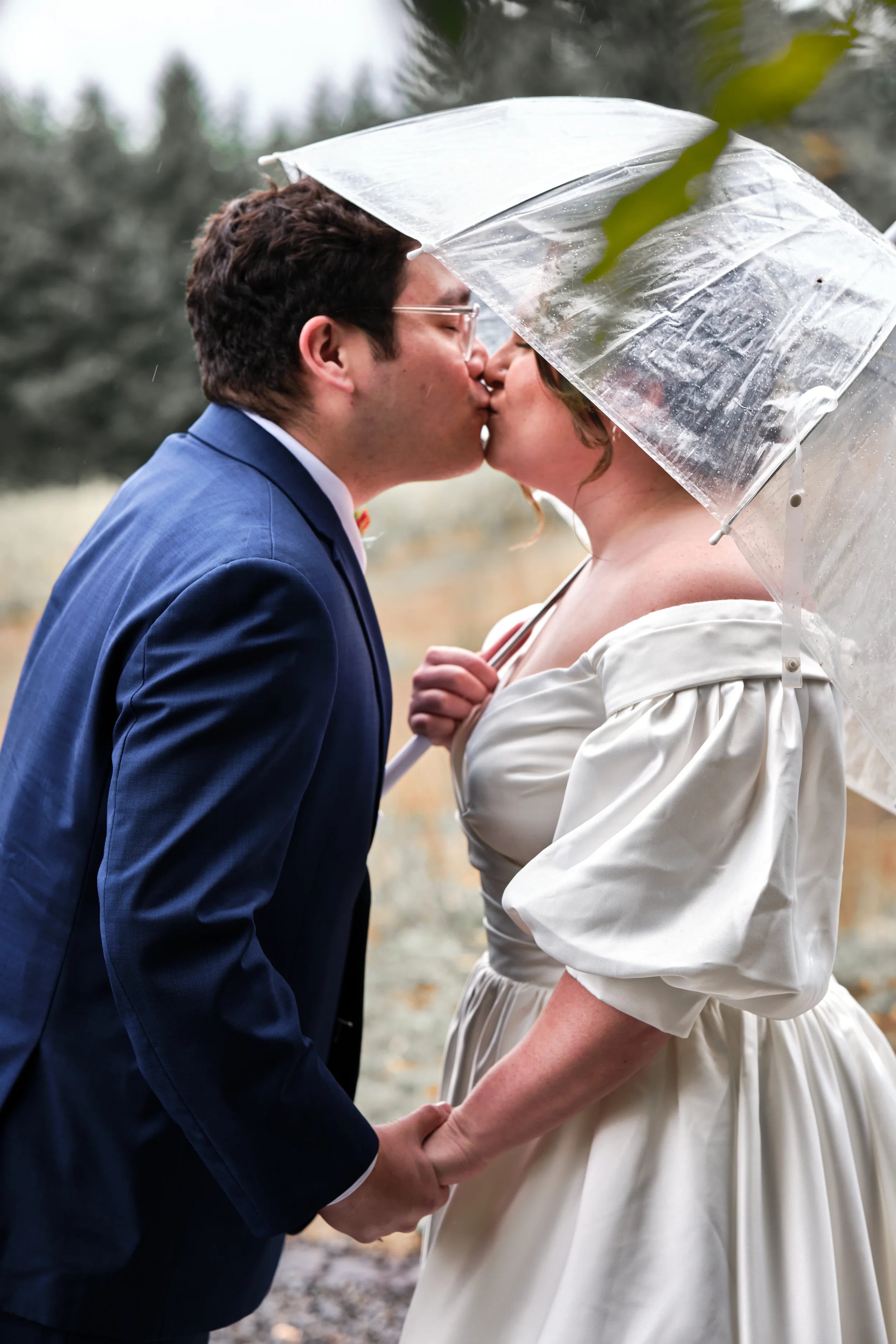 A couple kissing outdoors under a transparent umbrella, holding hands, with trees in the background during rainy weather.