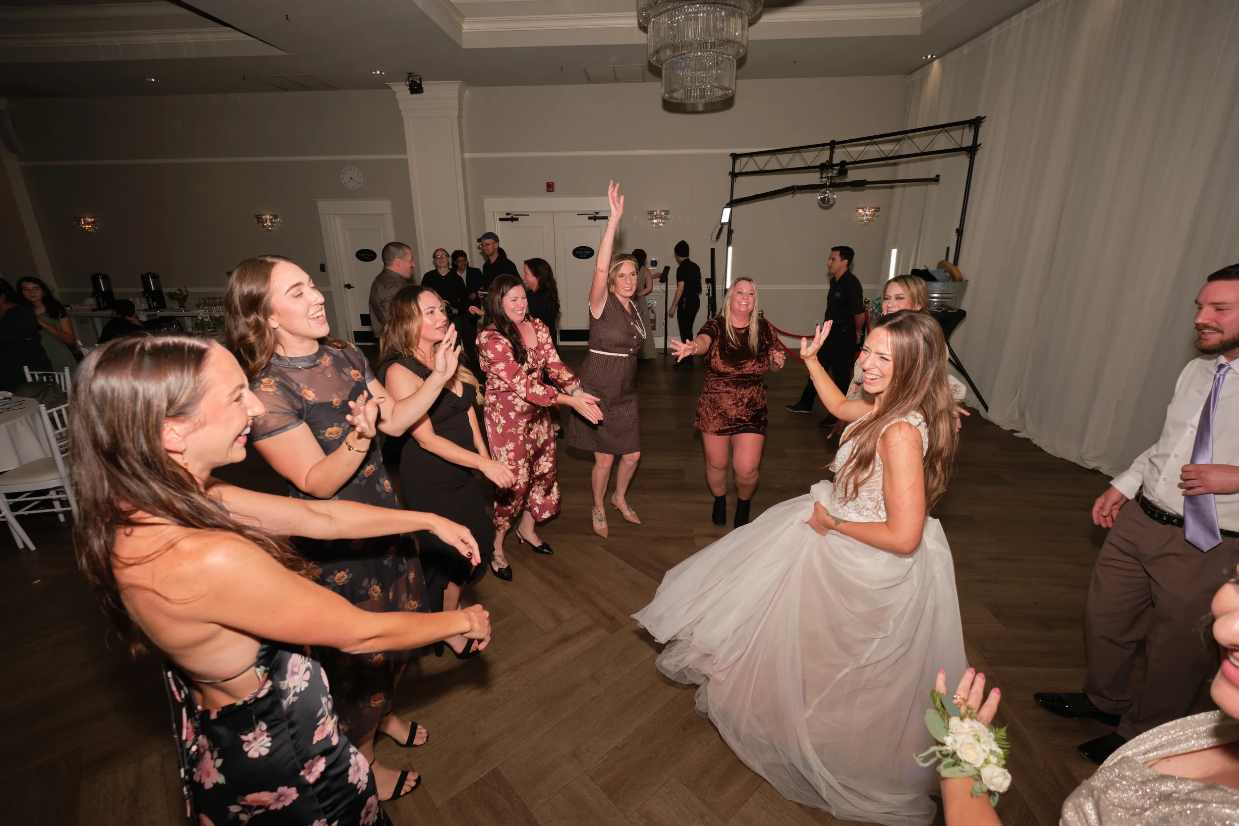 People dancing at a wedding reception. The bride, in a white wedding gown, is smiling and dancing with several guests. The guests, mostly women, are dressed in elegant dresses and are enjoying the celebration.