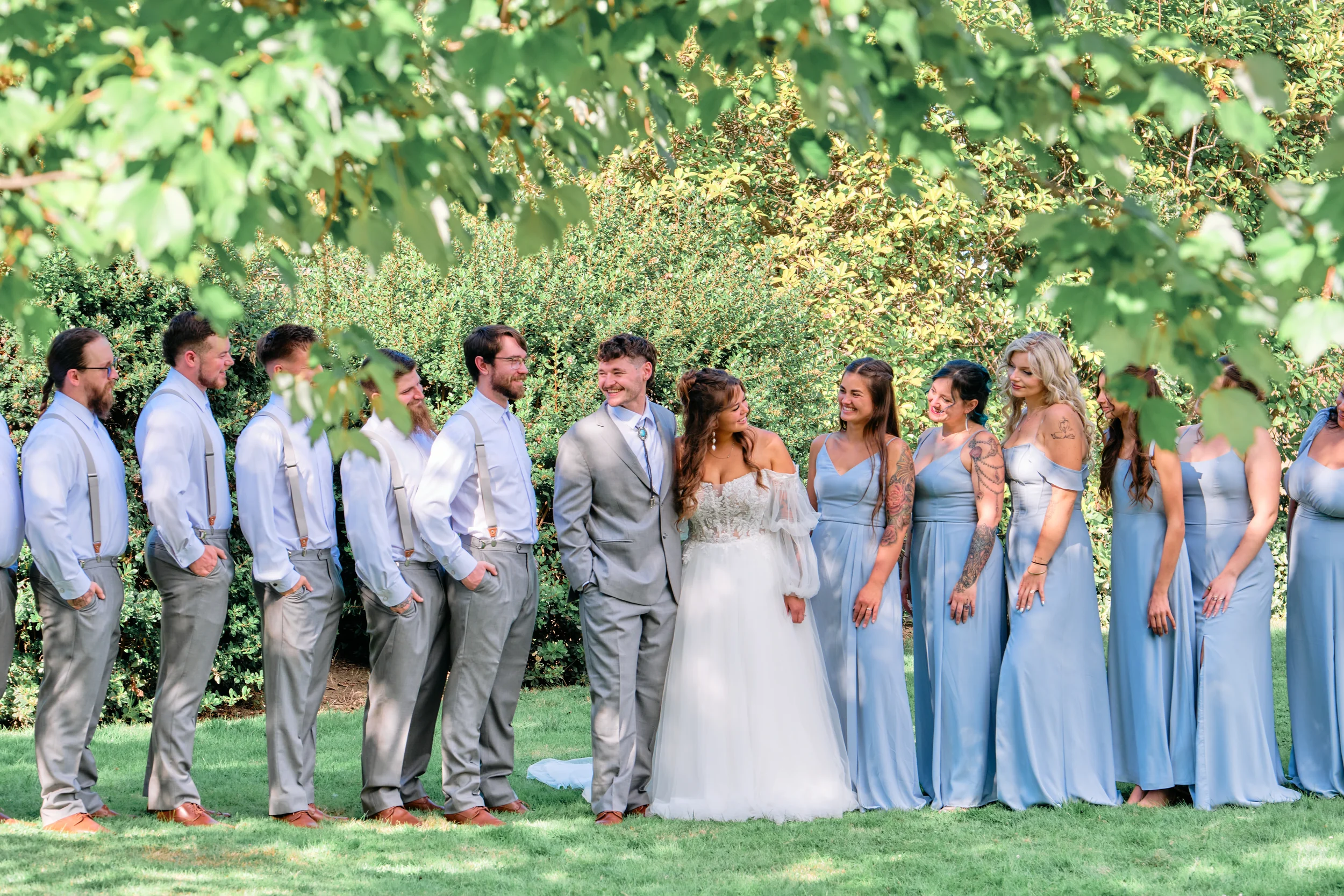 A wedding party standing outdoors in front of green leafy bushes, with the groom and bride in the center, surrounded by bridesmaids and groomsmen, all dressed in formal attire, smiling, during a sunny day.