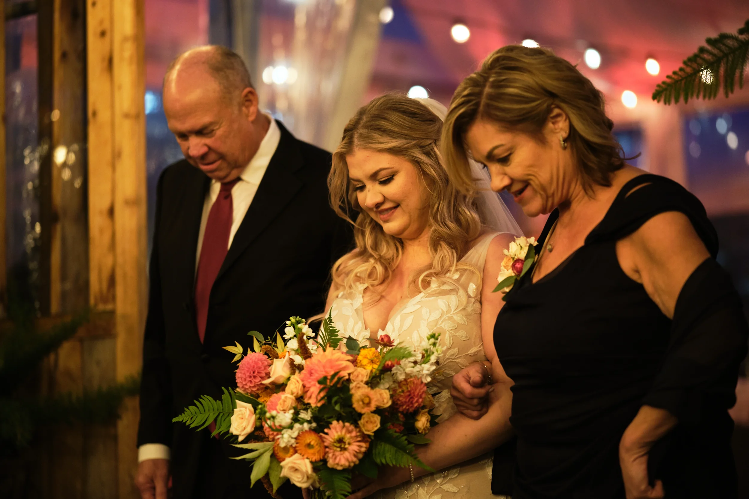 Three women and a man at a wedding, two women holding a bouquet of colorful flowers, all dressed formally and standing closely together, with a warm, festive background with blurred lights.