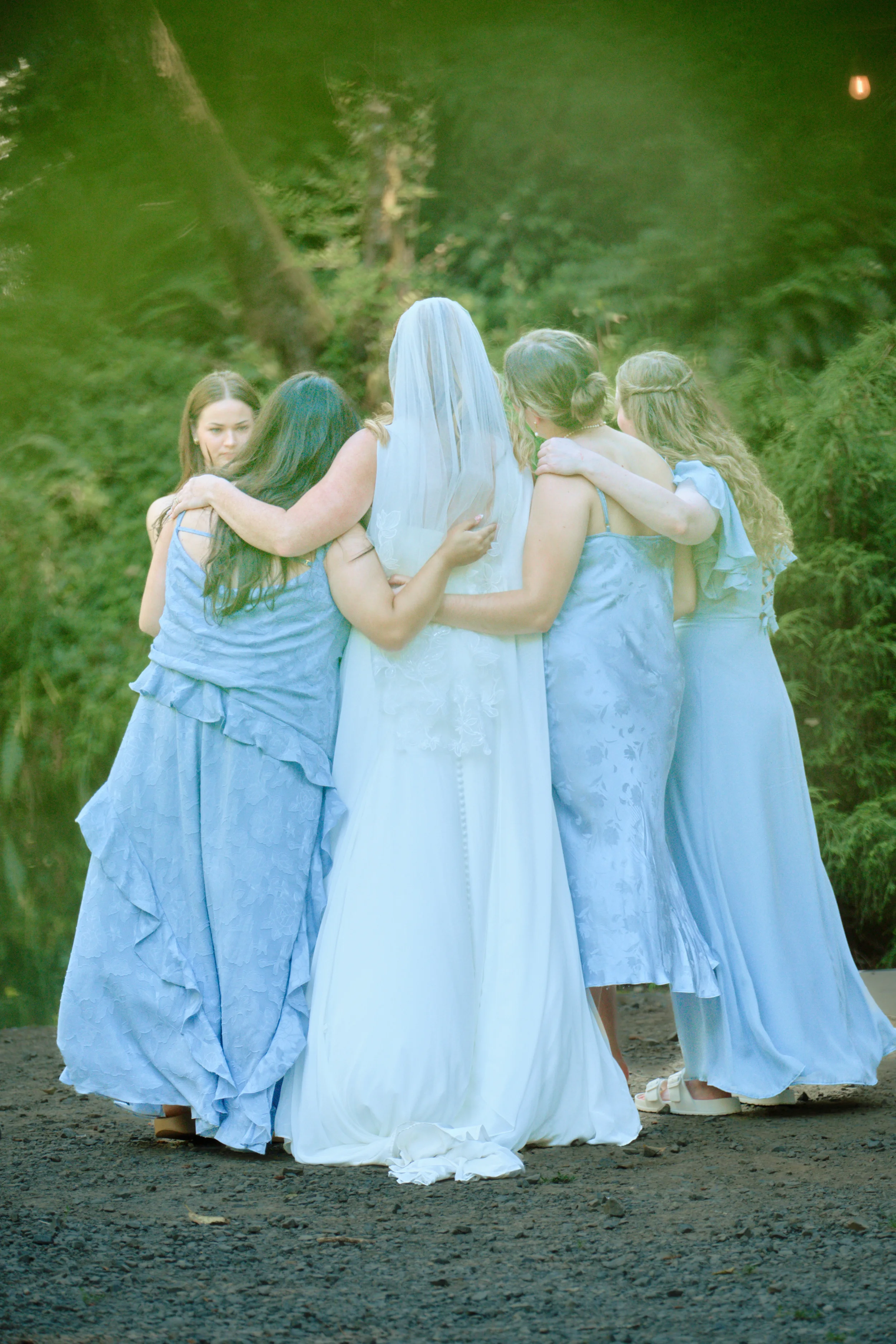 A group of women dressed in blue dresses and one woman dressed as a bride in a white gown and veil, embracing each other outdoors in a circle during a wedding ceremony.