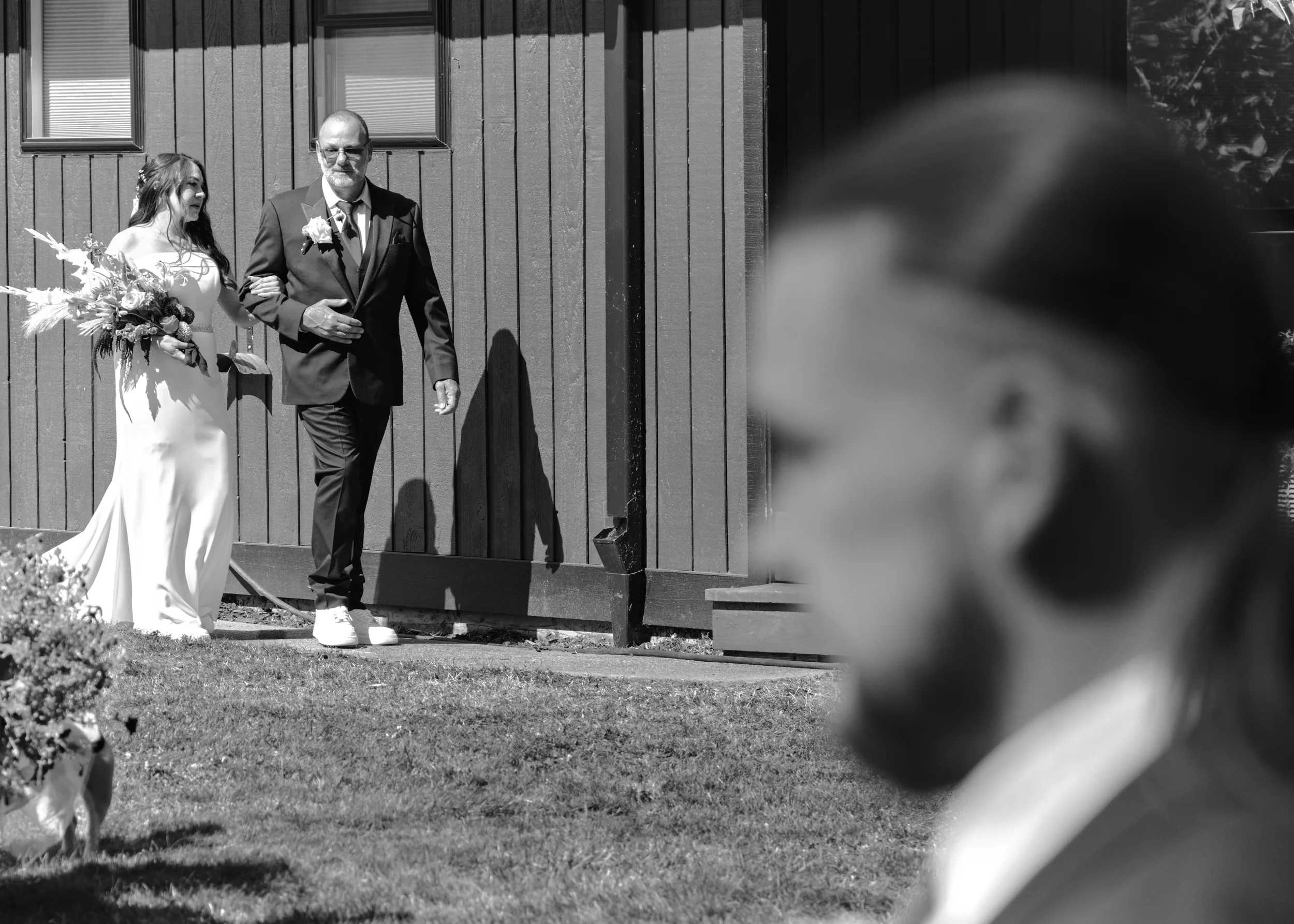 A bride and an older man, likely her father, walking arm in arm outside a building with vertical wooden siding, during a wedding ceremony. The bride carries a bouquet and wears a dress, while both are looking downward.