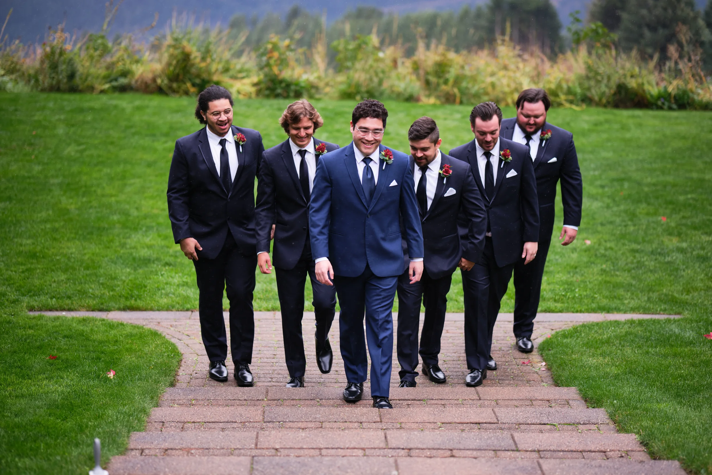 A group of six men dressed in dark suits, white shirts, and black shoes walking on a paved path in a lush green park or garden. They are smiling and appear to be happy, with some wearing boutonnières with red flowers on their jackets. The background 