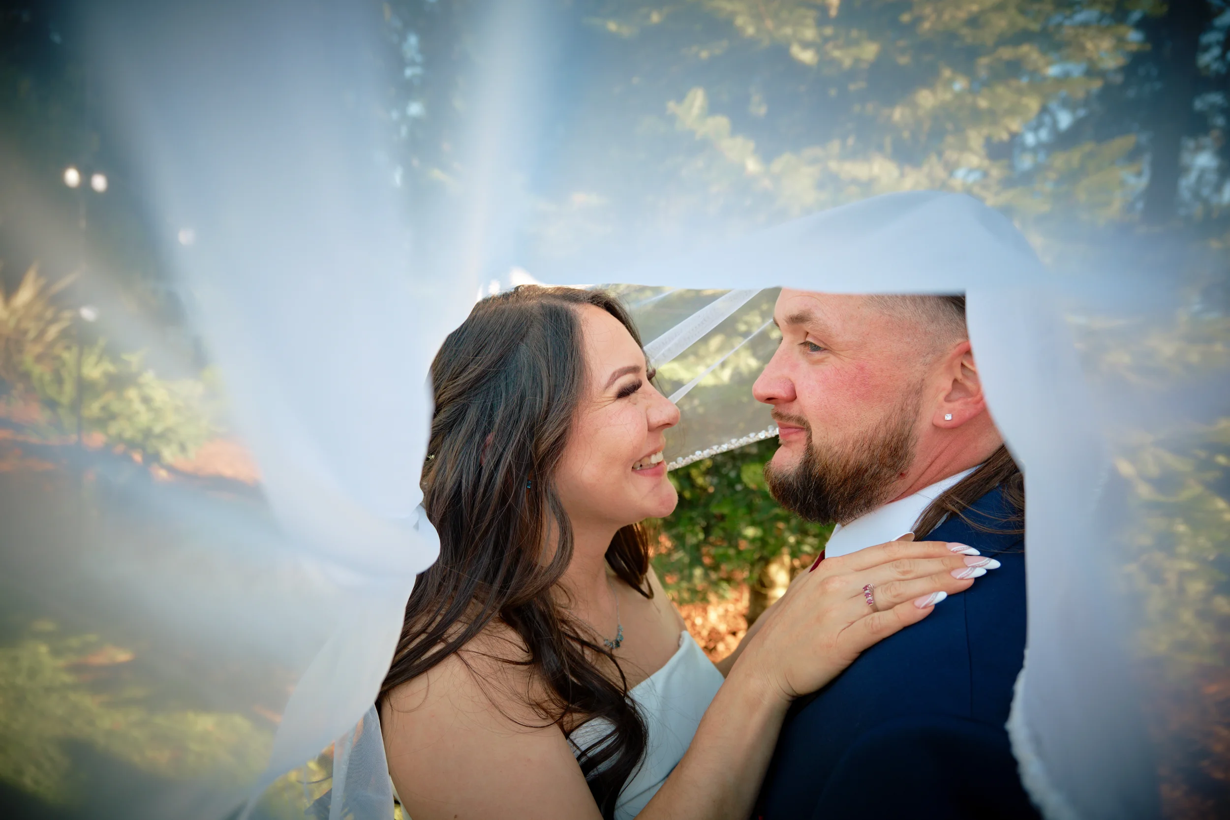 A bride and groom facing each other closely with smiles, inside a wedding veil that frames the scene, outdoors with trees in the background.