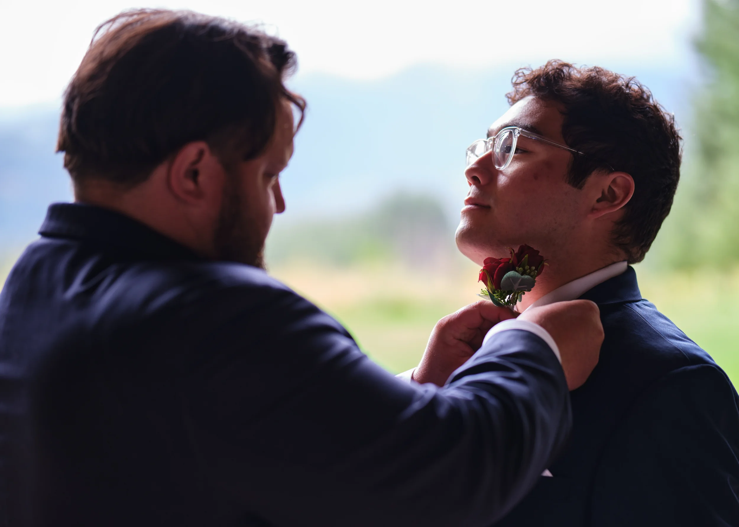 Two men, one with glasses, are in close proximity, with one man placing a flower bouquet near the other's neck in an outdoor setting with blurred greenery background.