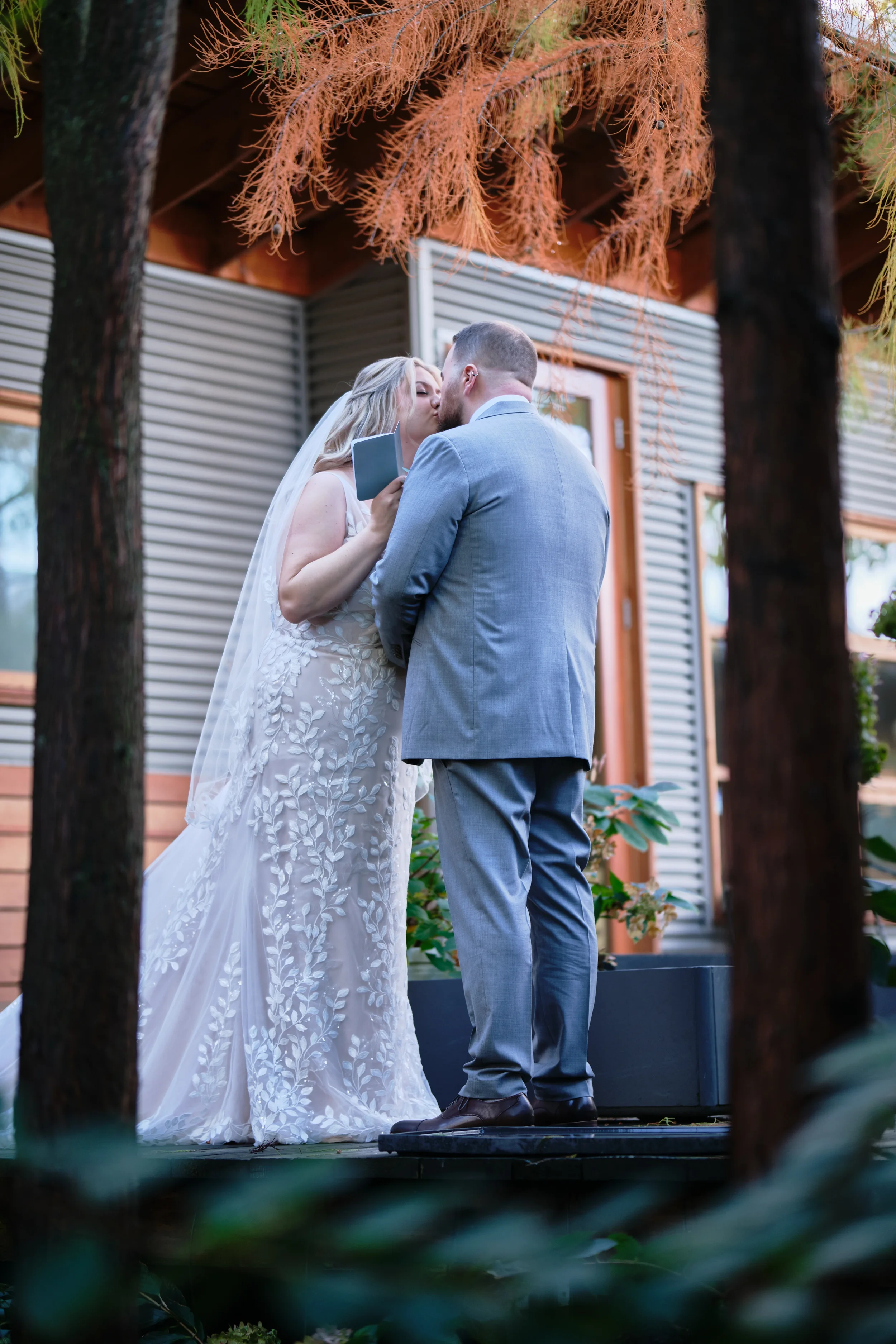 A bride and groom kiss during their wedding ceremony outdoors, surrounded by trees and nature.