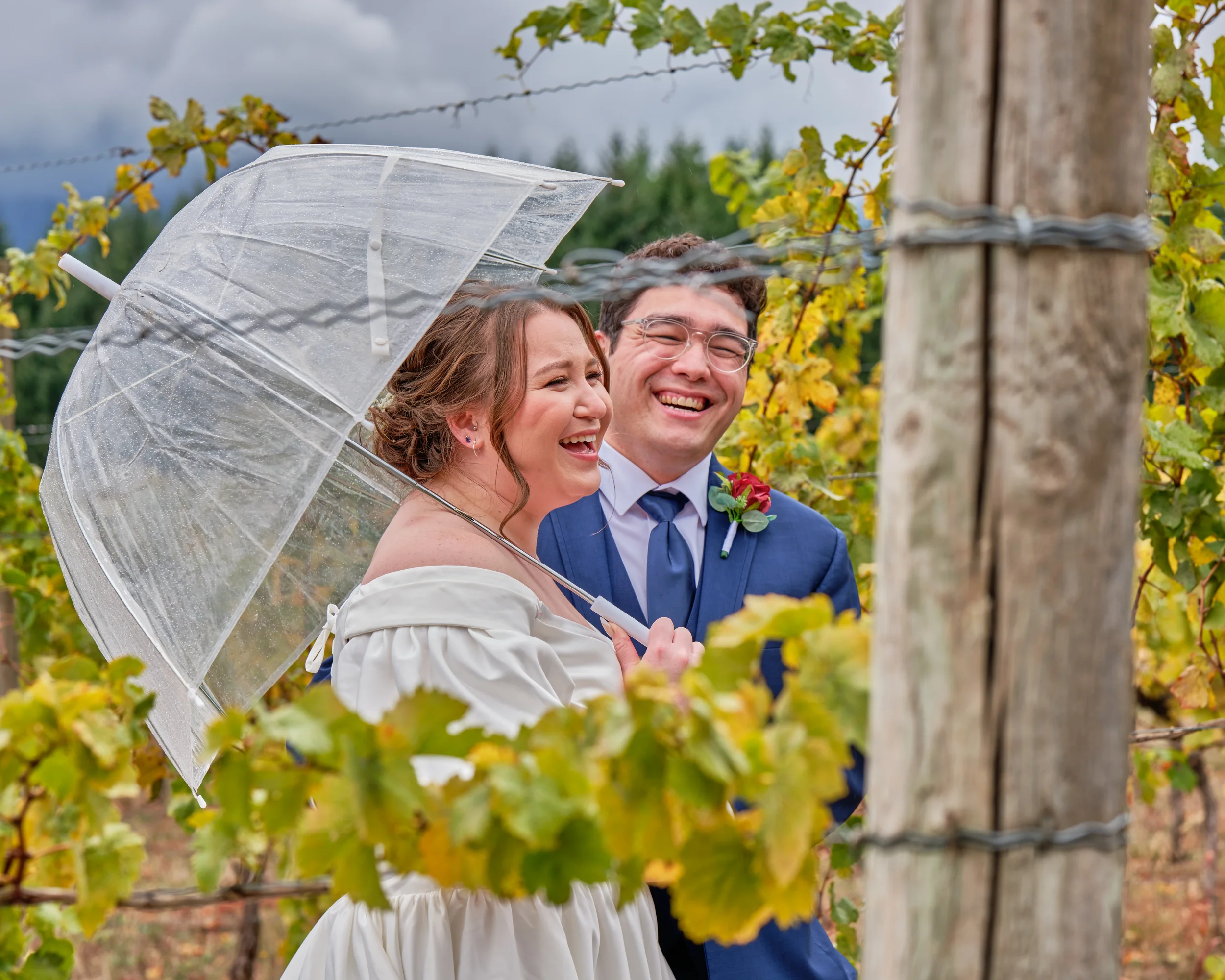 A happy couple at a vineyard, with the woman holding a clear umbrella. They are smiling and dressed in formal attire, suggesting a special occasion like a wedding.