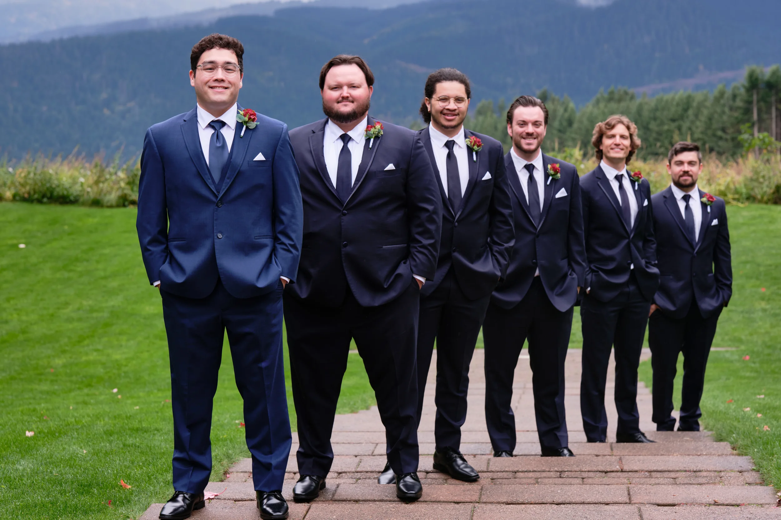 Group of six men wearing formal suits with boutonnières, standing outdoors on a paved path with a grassy area and mountains in the background.