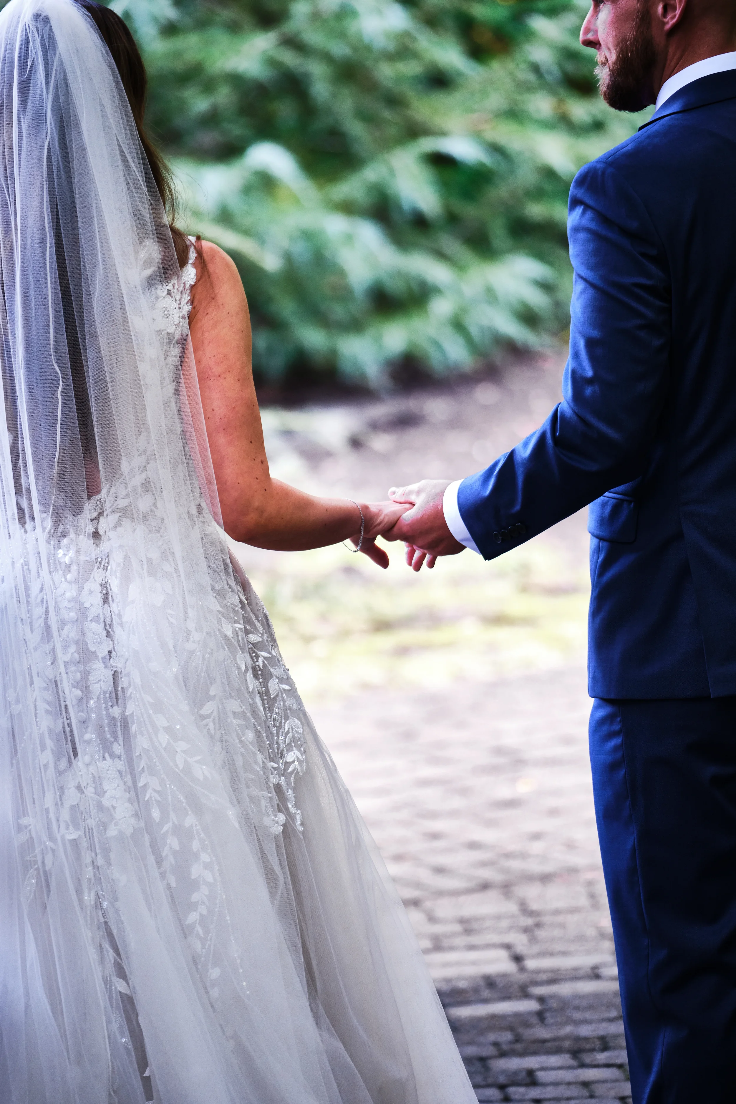 A bride and groom holding hands during their wedding ceremony outdoors, with the bride wearing a lace wedding gown and veil, and the groom in a navy suit, standing on a stone pathway surrounded by greenery.