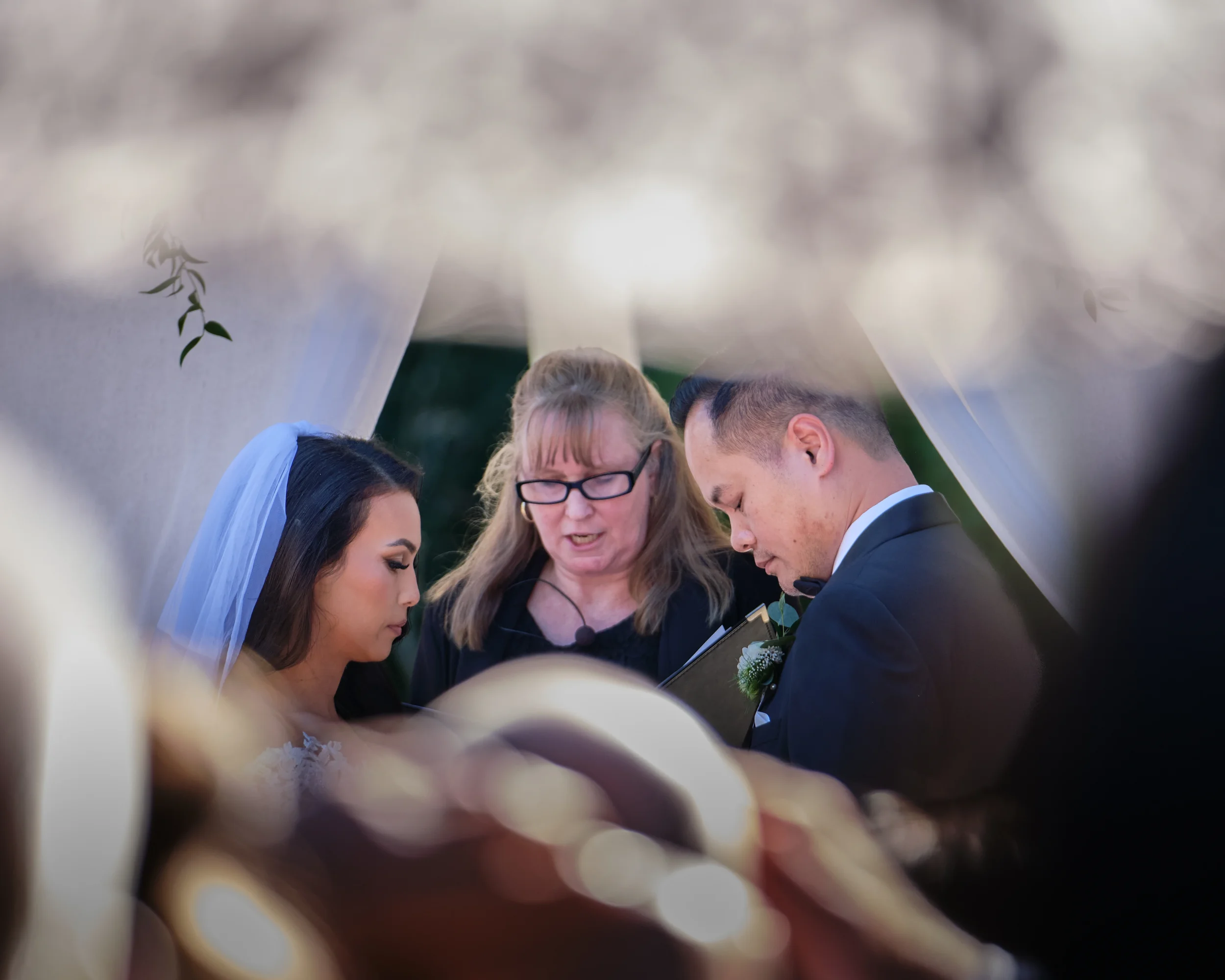 A group of people, including a bride and groom, are standing closely together during a wedding ceremony. An officiant is in the center, reciting the vows or prayers, with their heads bowed. The scene is outdoors with greenery in the background.