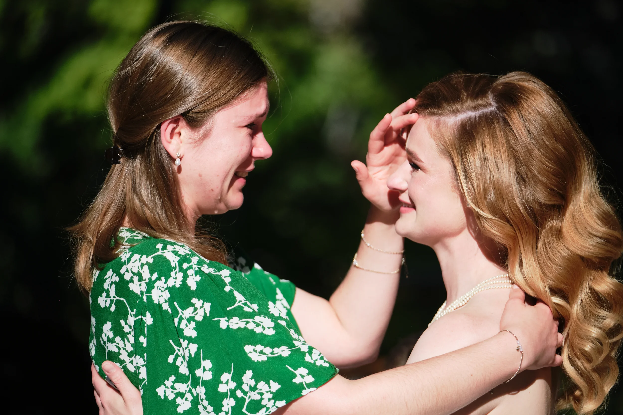 Two women smiling at each other, holding each other's faces gently, outdoors at night with trees in the background.