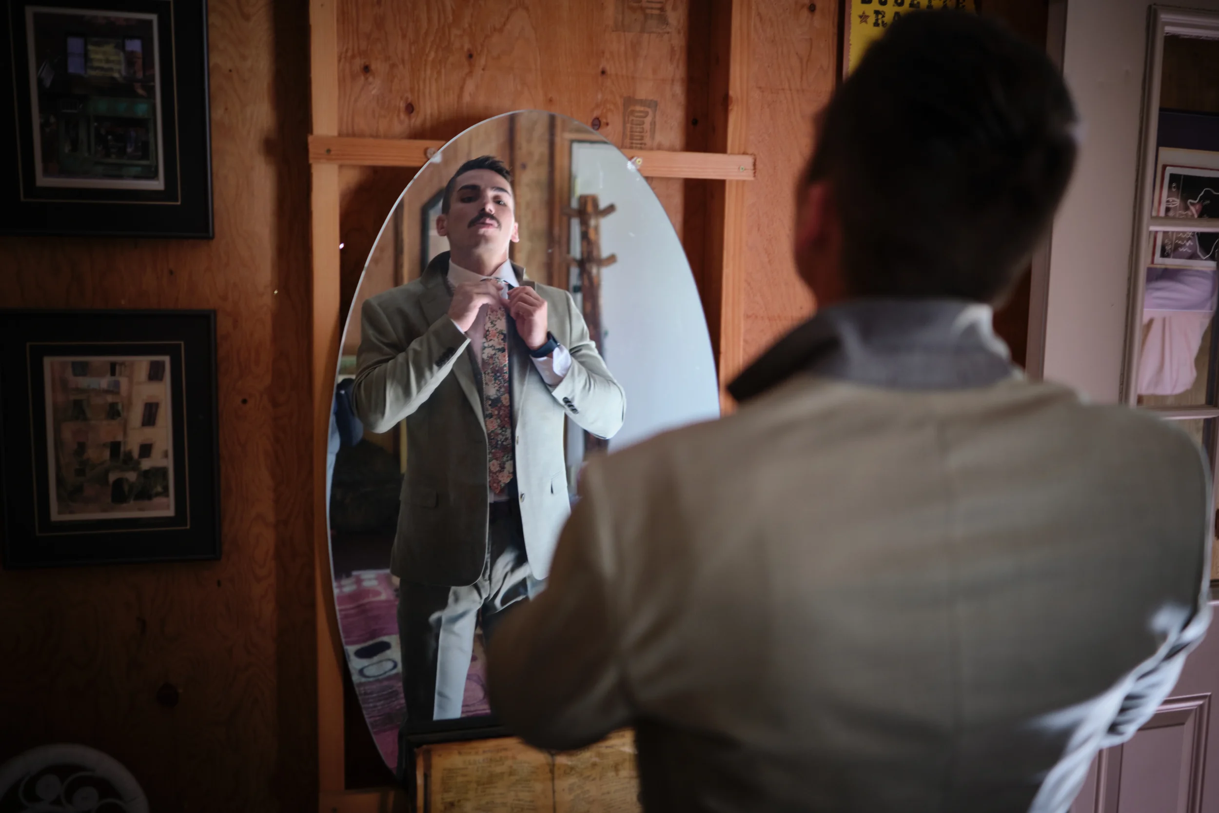 A young man adjusting his tie while looking at himself in an oval mirror in a wooden-paneled room.