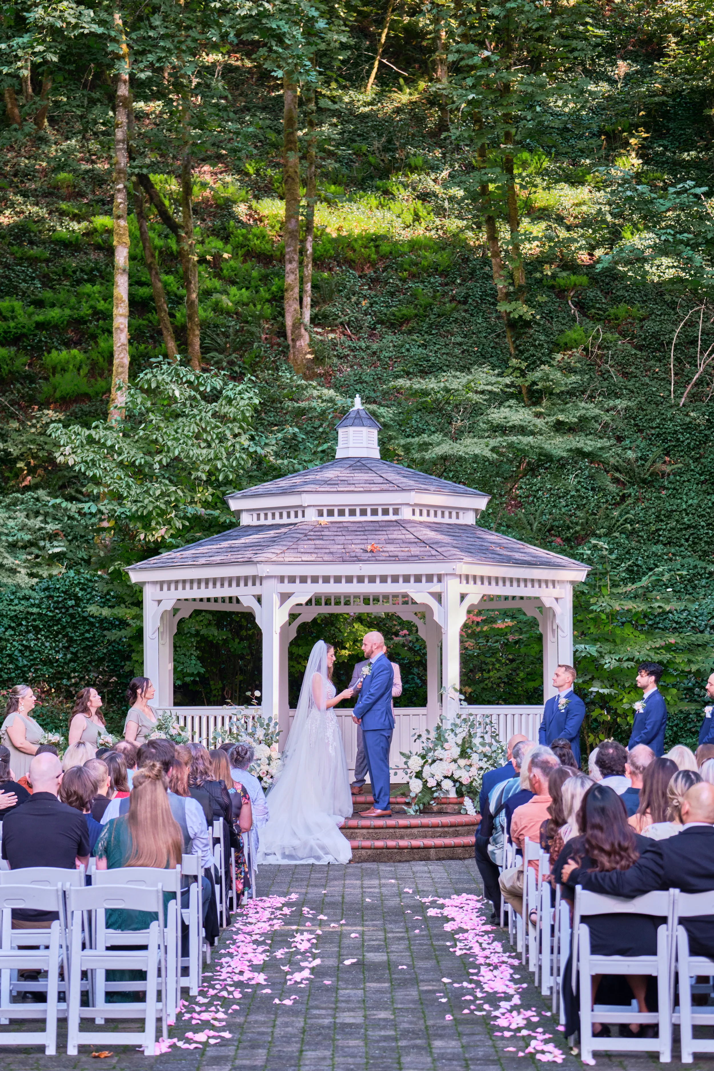 A couple getting married outdoors under a white gazebo surrounded by guests, with a forested backdrop and pink flower petals scattered on the aisle.